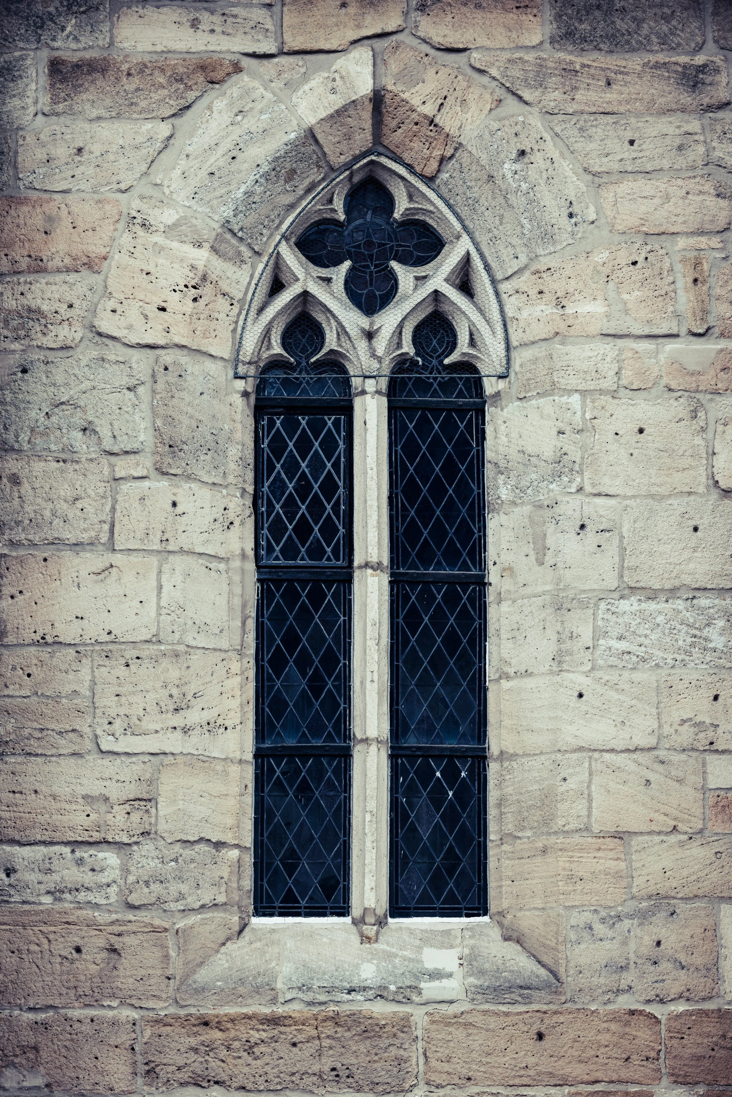 A stained glass window with a Gothic design, featuring a large four-petal flower pattern at the top and smaller leaf-like shapes below, set in a stone wall.
