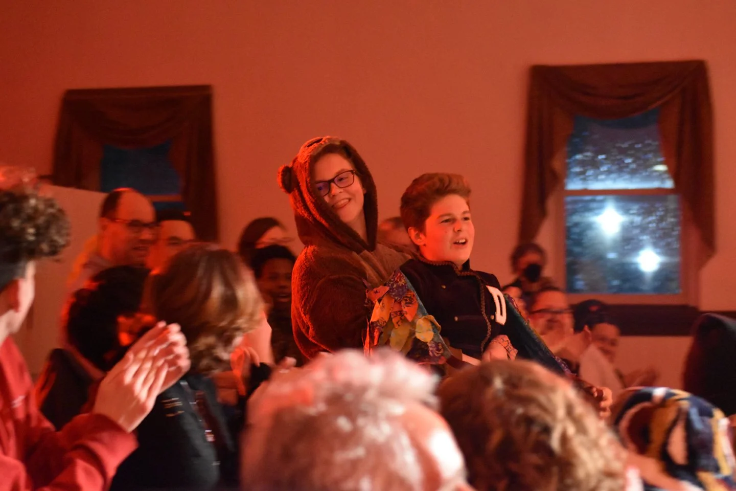 People gathered in a room, watching and clapping for a performer. Two young individuals are standing, smiling, and enjoying the show, with people around them clapping and cheering.