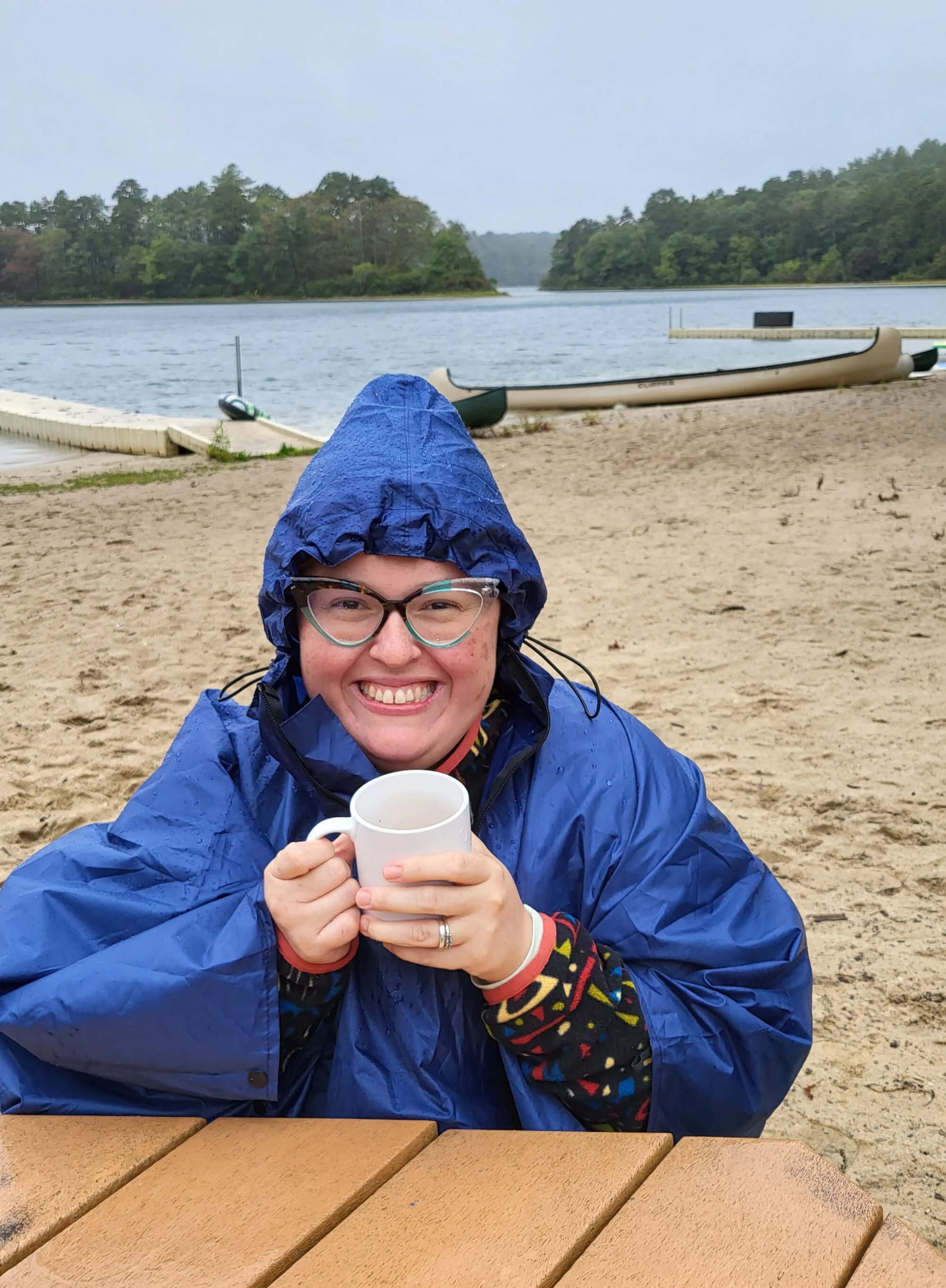 Person with glasses in a blue rain jacket smiling and holding a white mug on a sandy lakeside beach, overcast sky, trees across the water, a canoe and a dock are visible in the background.