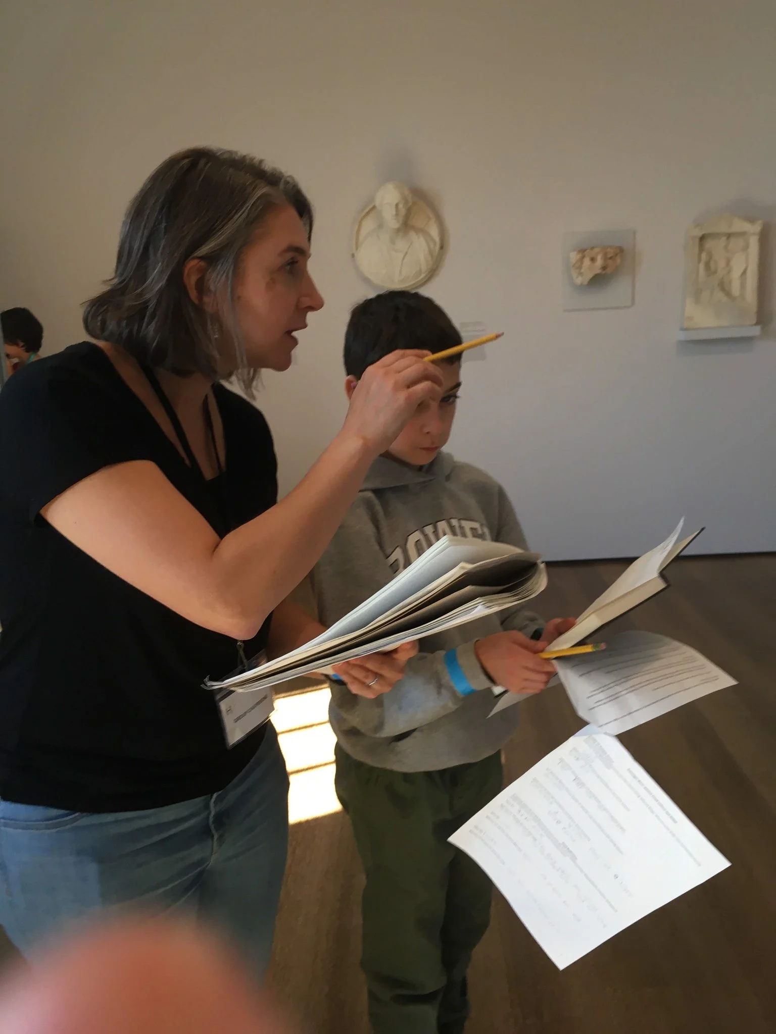 A woman with shoulder-length gray hair and a young boy, both holding papers and books, standing at a table with additional papers, in an art gallery with wall-mounted sculptures.