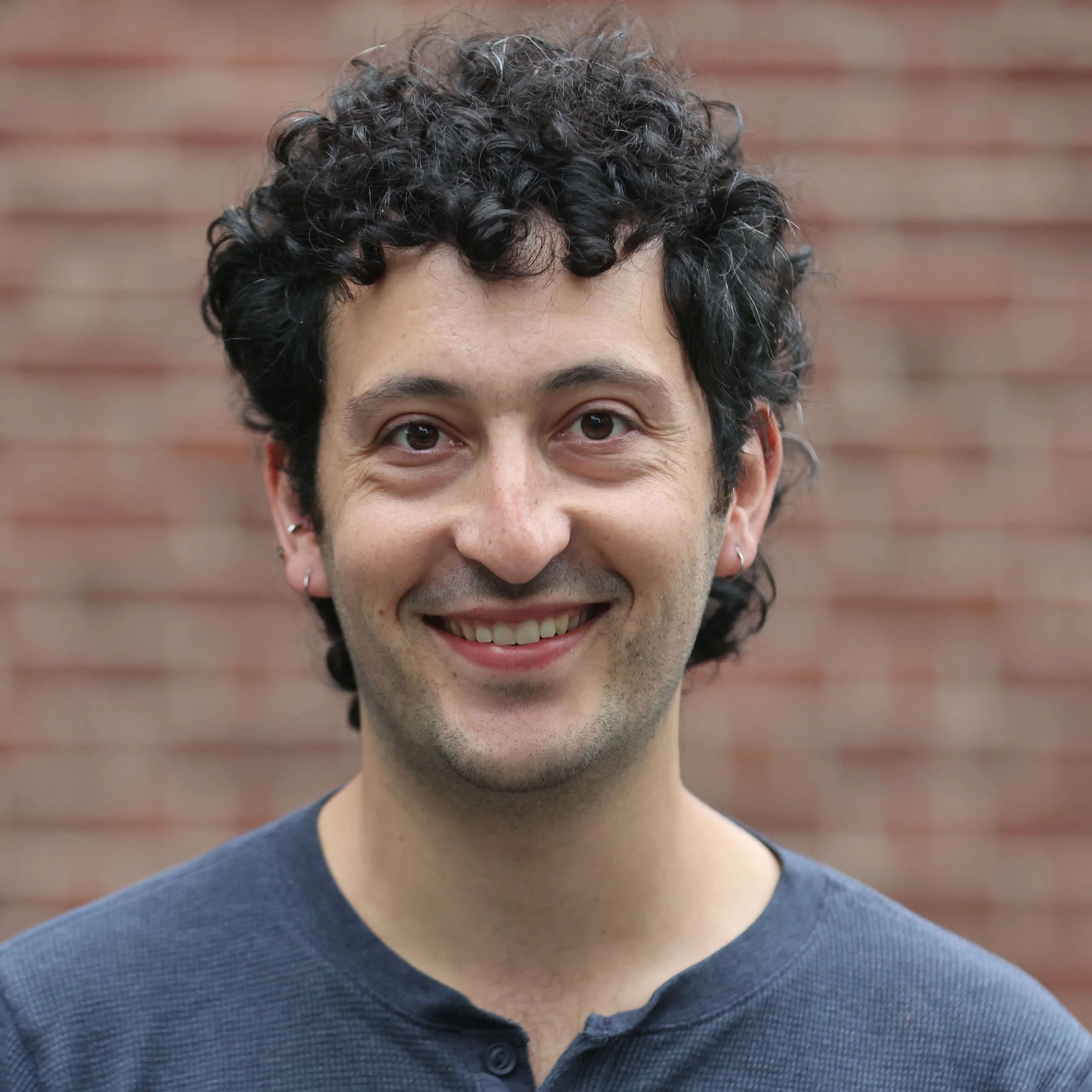 Close-up of a person with curly dark hair, brown eyes, smiling, wearing a dark gray button-up shirt against a blurred brick wall background.