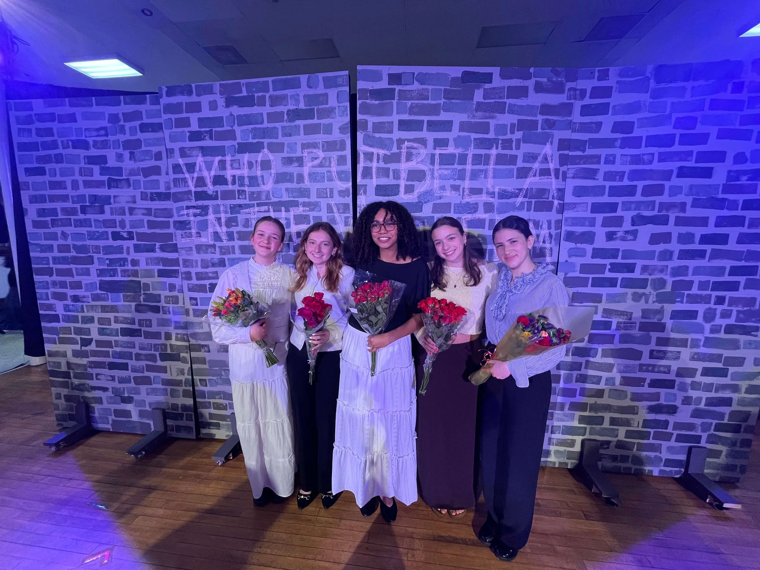 Five women standing in front of a brick-patterned wall with the words "MARCH REBEL" written on it, holding bouquets of flowers, under purple lighting.