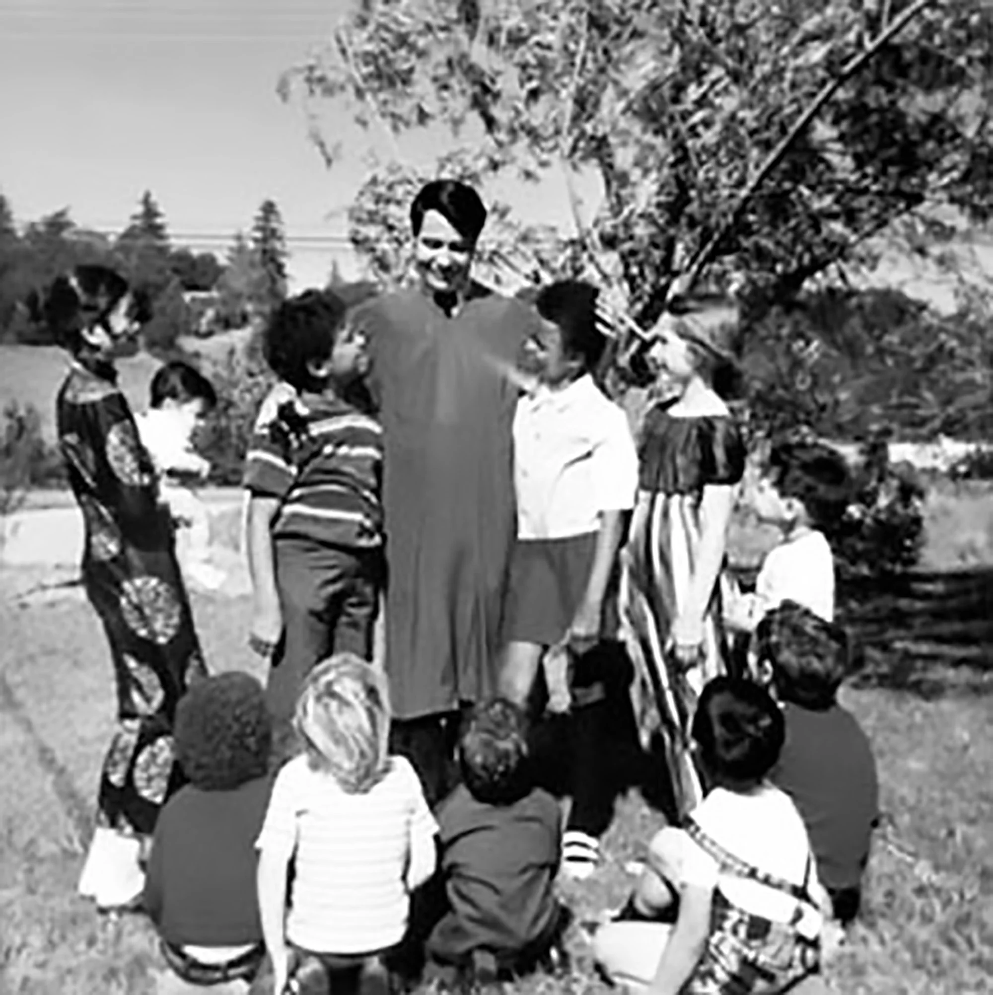 A black and white photo of four children gathered outdoors on a sunny day, standing near a tree, with houses or buildings in the background.