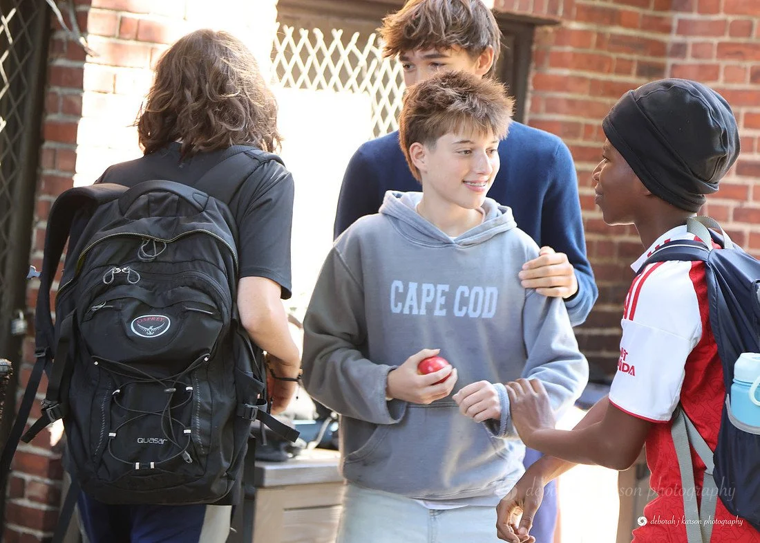 Four teenagers having a chat outside a brick building, one with a backpack, holding an apple, wearing a gray hoodie that says 'Cape Cod', and another wearing a black head covering and a red and white sports jersey.