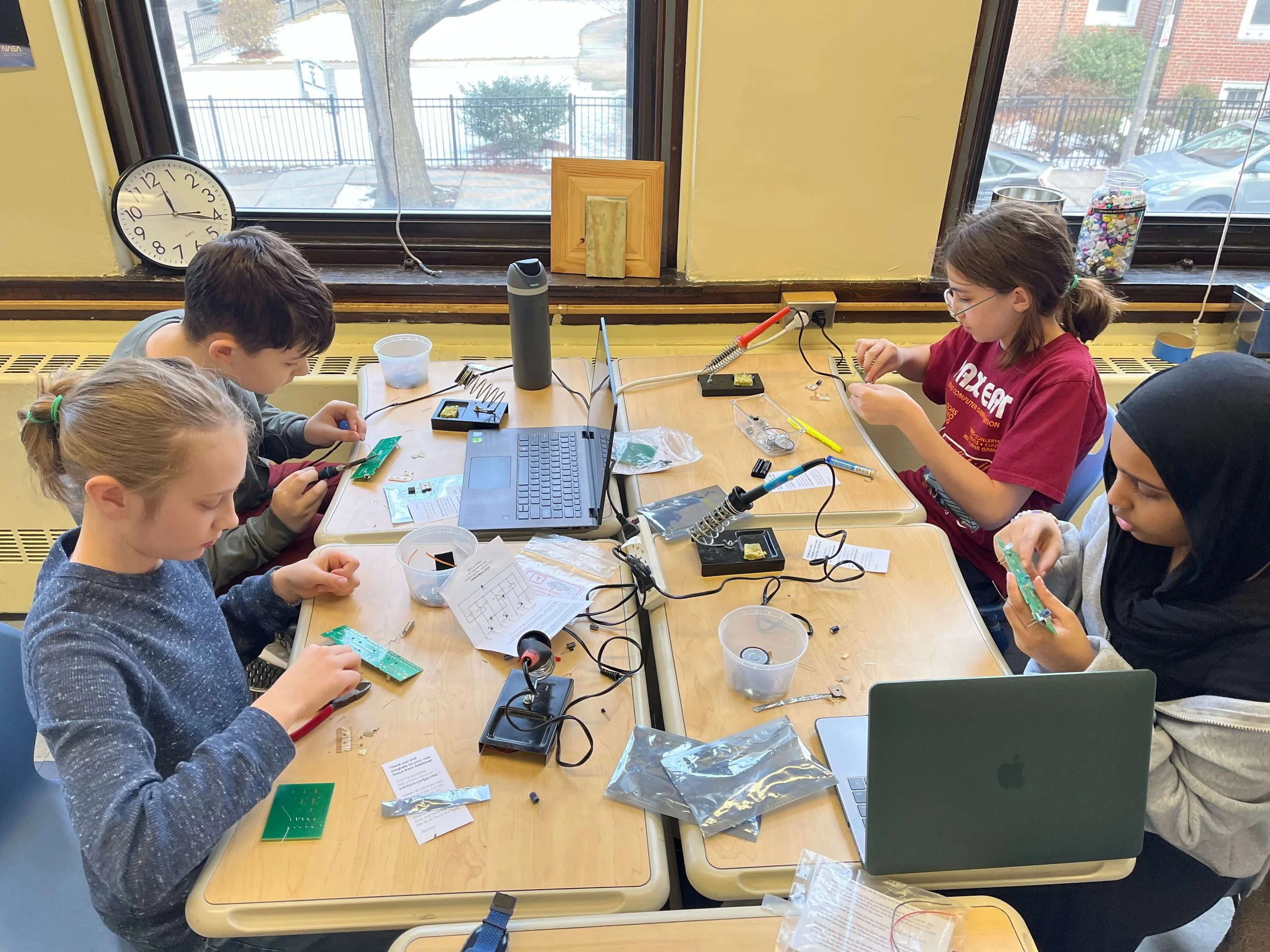 Children working on electronics projects at a table with tools, wires, and circuit boards in a classroom near a window.