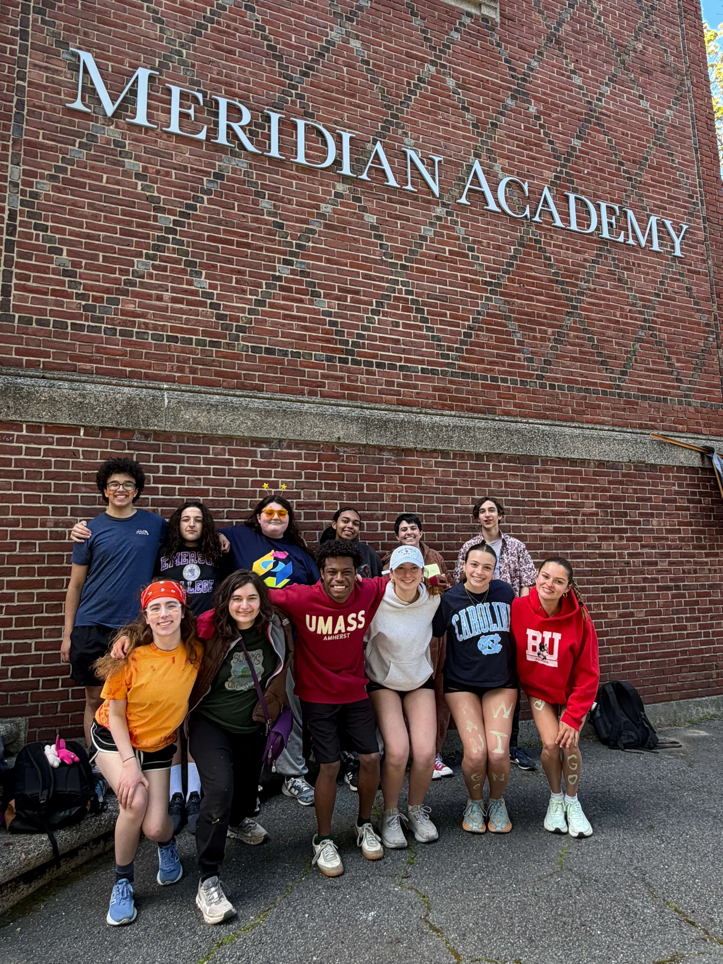 Group of young people standing in front of a brick wall with the words 'Meridian Academy' on it. They are smiling and appear to be on a school trip or outdoor activity, some are wearing athletic clothing and backpacks.