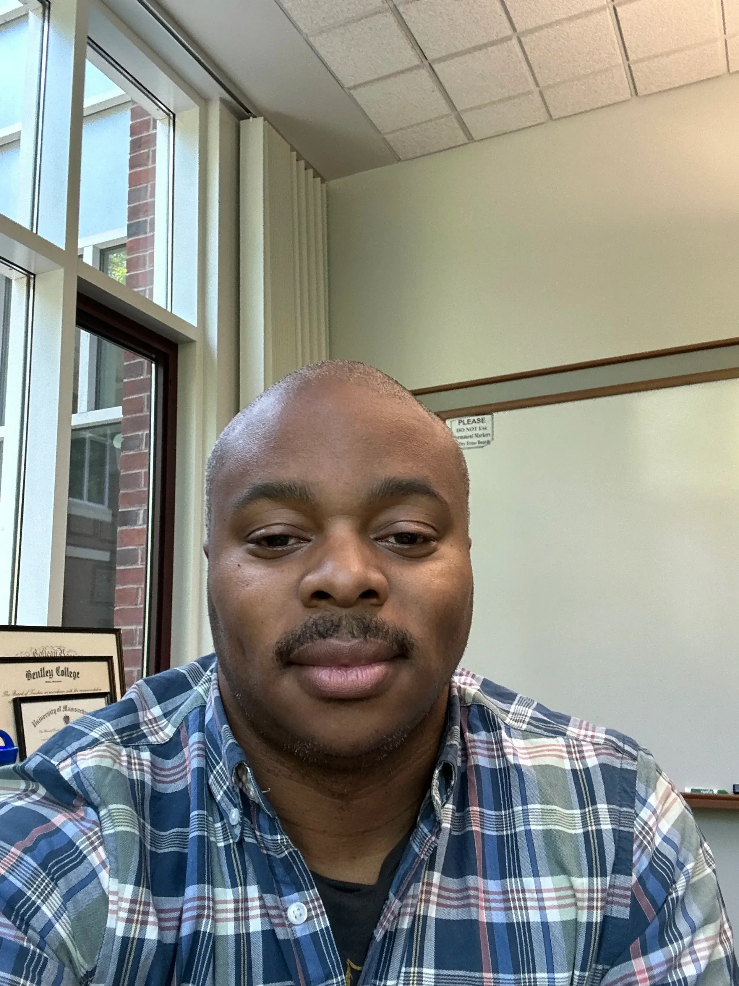 A man with a dark complexion and a shaved head, wearing a multicolored plaid shirt, sitting indoors near a window with beige walls and signs, with part of his face and shoulders visible.