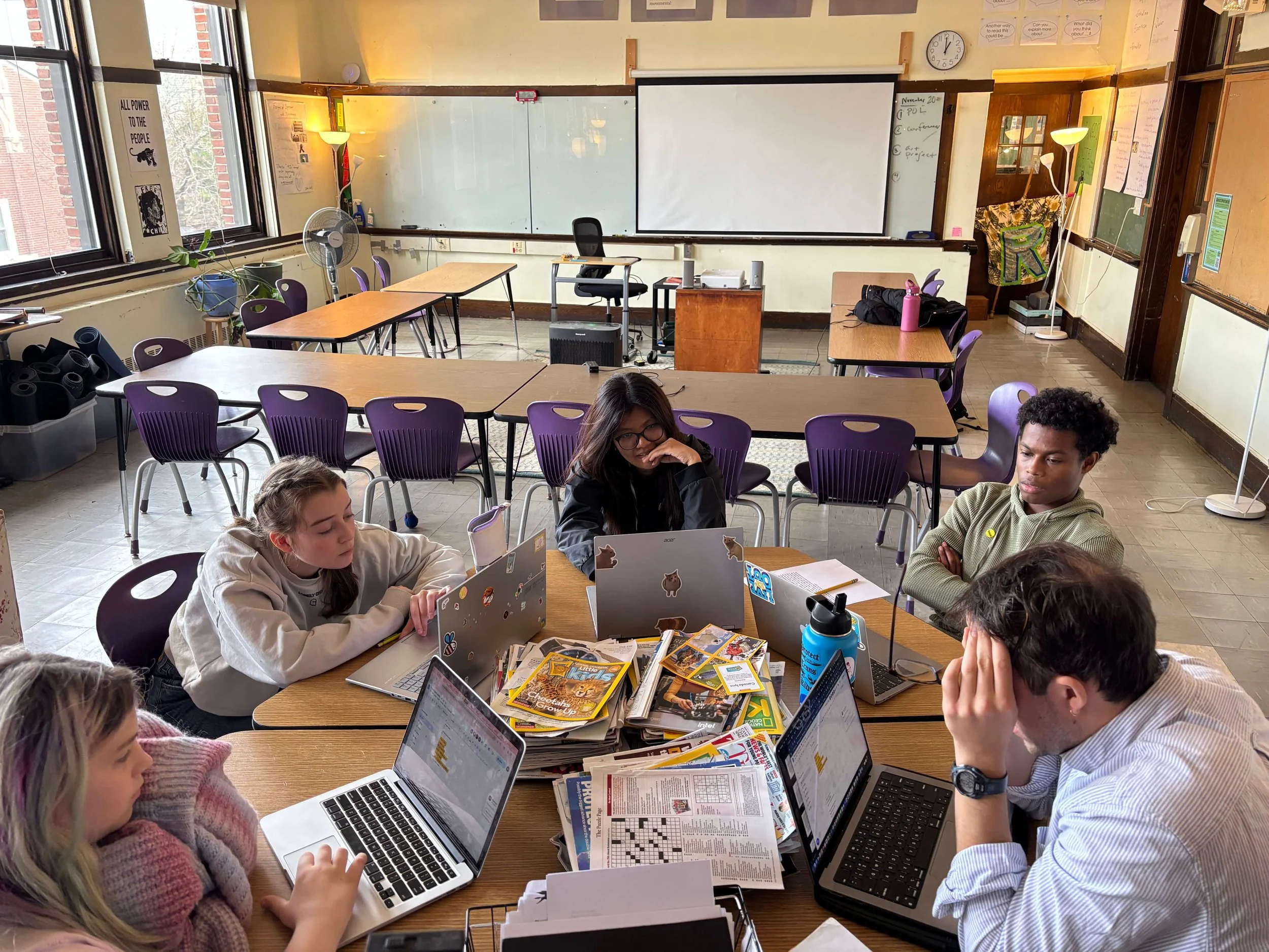 A classroom with students working on laptops at tables, some with open books and magazines, in front of a whiteboard and projector screen.