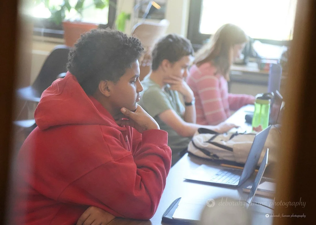 Three students sitting at a desk in a classroom, focused on their laptops, with a green water bottle and backpack nearby.
