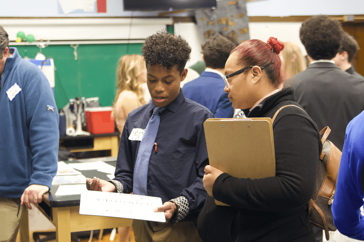 A young man wearing a blue shirt and tie holding a whiteboard, talking to a woman with red hair and glasses holding a clipboard, among other people in a busy indoor environment.
