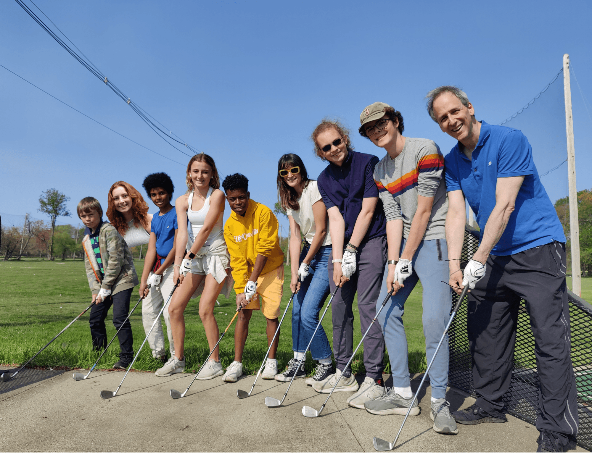 A diverse group of people standing in a line on a golf course, holding golf clubs, preparing to tee off, with a clear blue sky and trees in the background.
