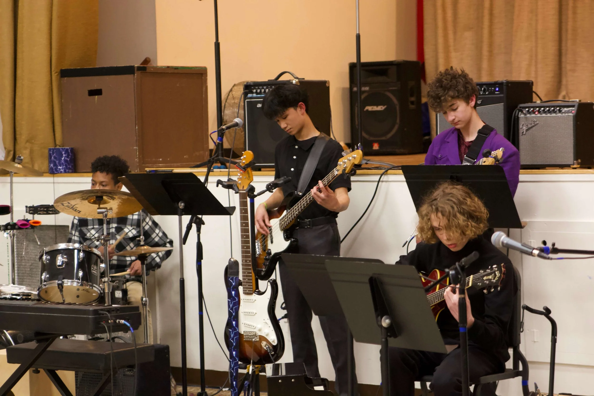 Young musicians performing with instruments including drums, guitar, bass, and keyboard, on stage with amplifiers and music stands.