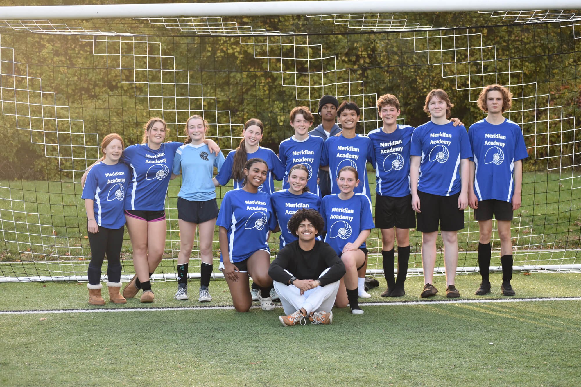 A group of young female and male soccer players in blue jerseys with 'Meridian Academy' written on them, posing behind a soccer goal on a grassy field with trees in the background.
