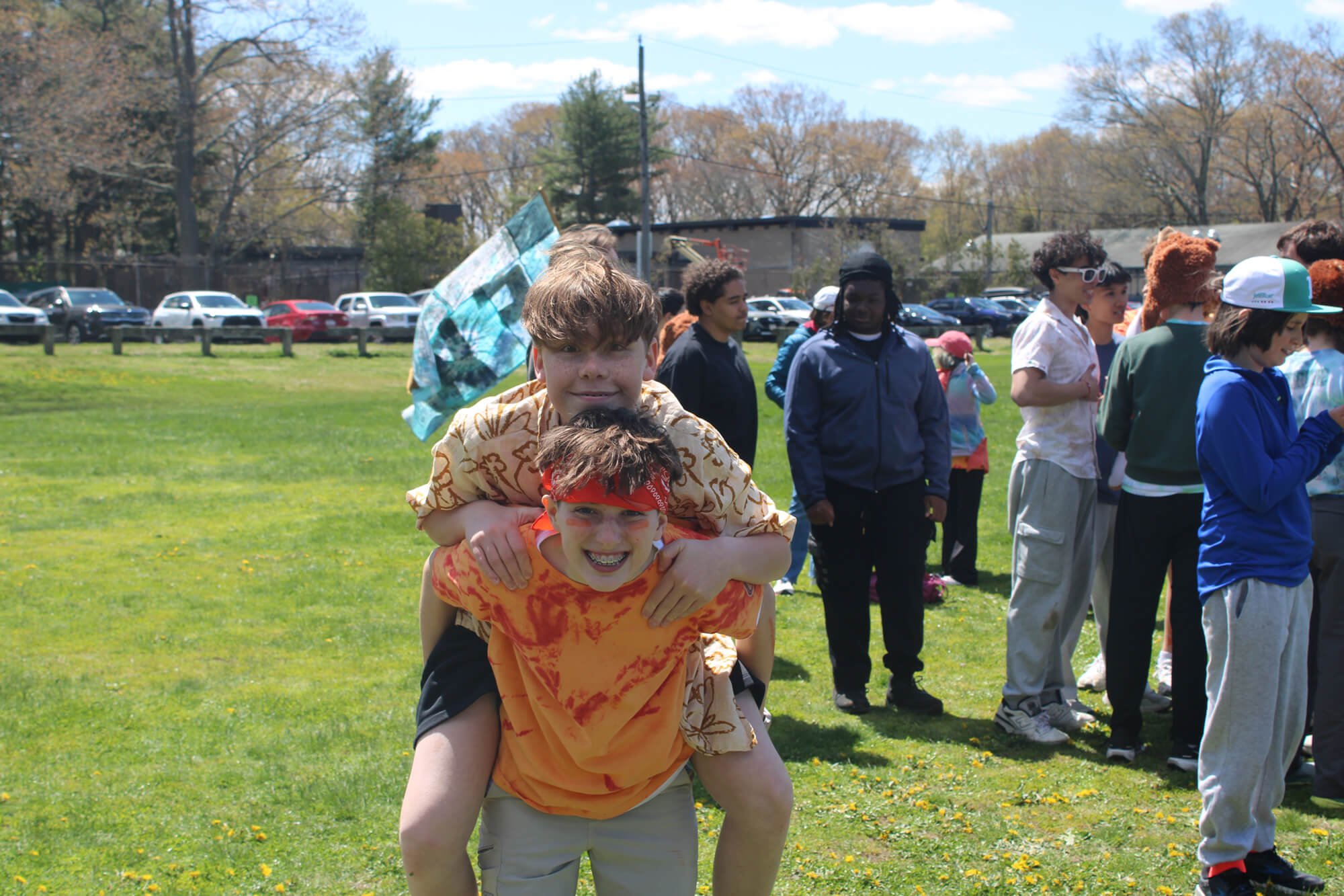 Children participating in an outdoor event, with some children on each other's shoulders, smiling and enjoying the sunny day on a grassy field with parked cars and trees in the background.