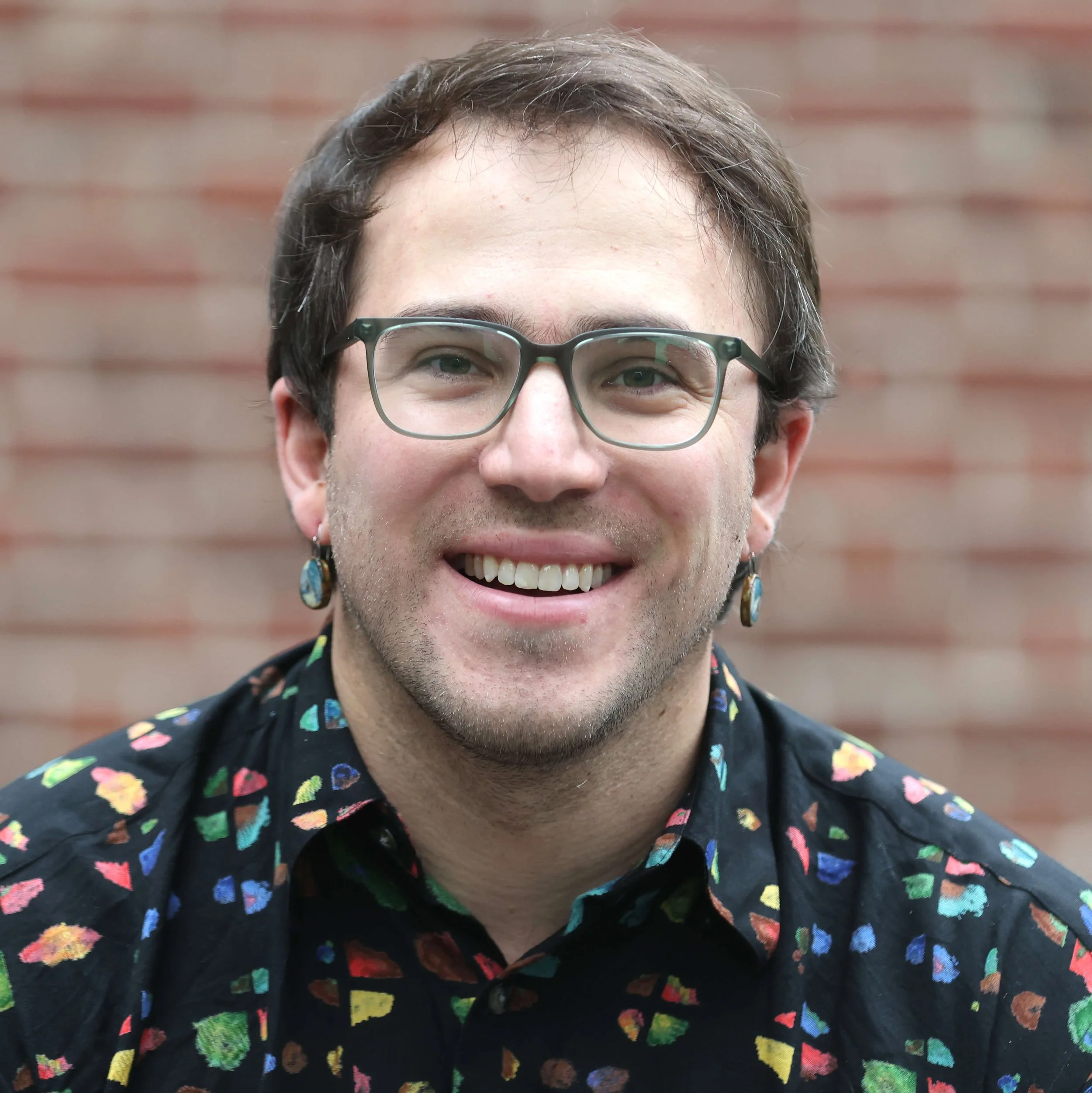A person with short dark hair, wearing glasses with a dark frame, and a black shirt with a colorful heart pattern, smiling in front of a blurred brick wall background.