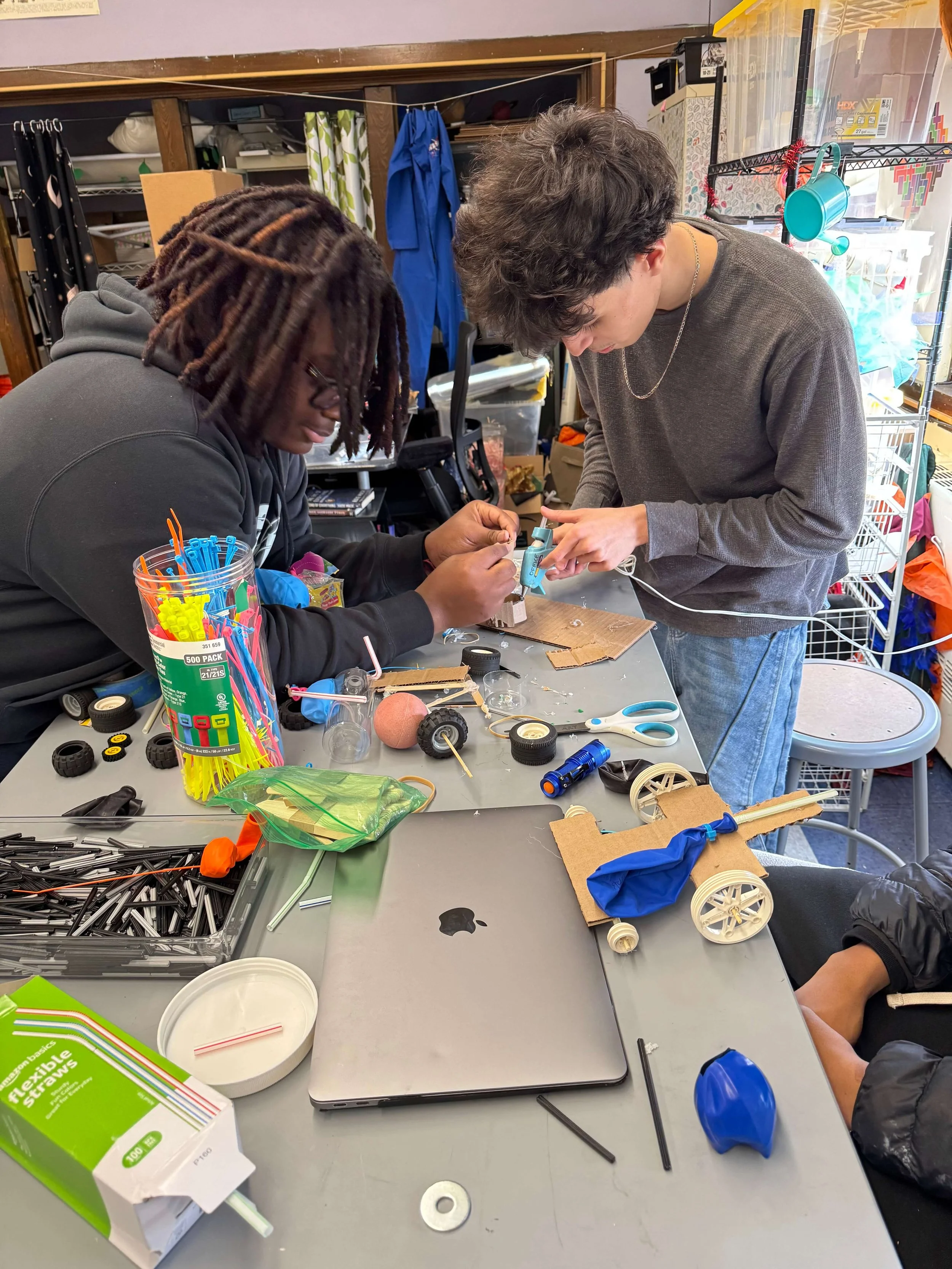 Two people working on a project at a cluttered table, assembling a small wheeled object surrounded by various tools and supplies in a workshop or classroom setting.