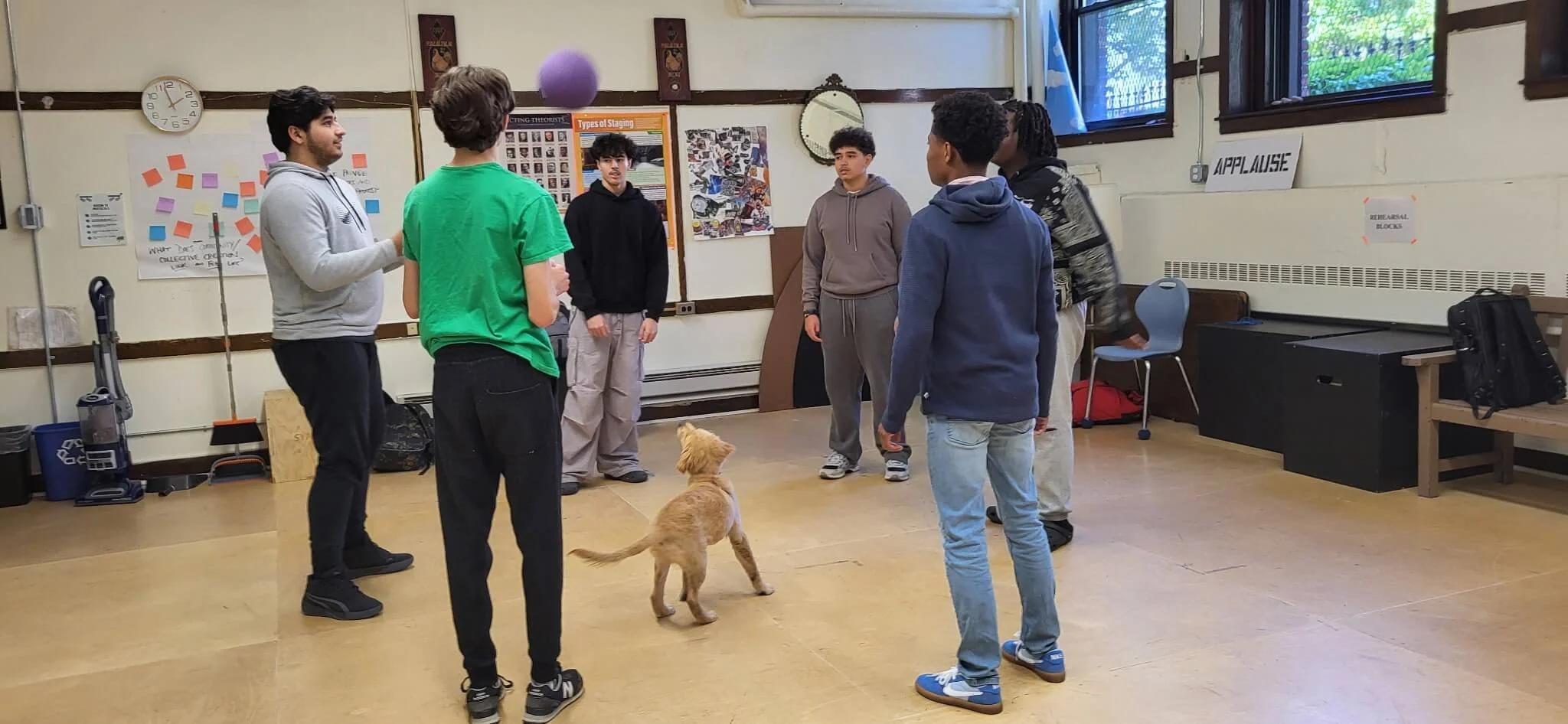 A group of young people standing in a circle with a dog in a classroom. One person is holding a purple ball. The classroom has bulletin boards, a clock, and windows, with signs on the wall that say 'APPLAUSE' and 'REUSABLE BLOCKS'.