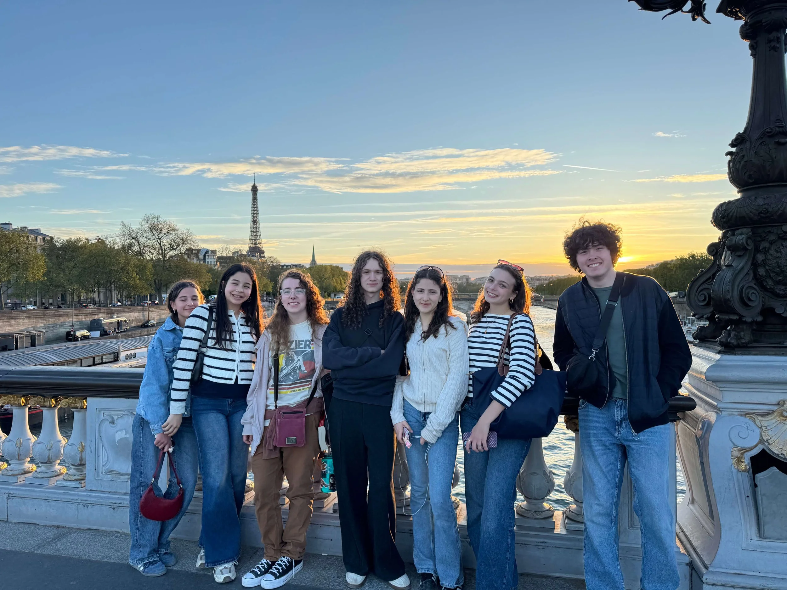 A group of seven young people standing together on a Paris bridge during sunset, with the Eiffel Tower visible in the background and a large ornate lamp post on the right.