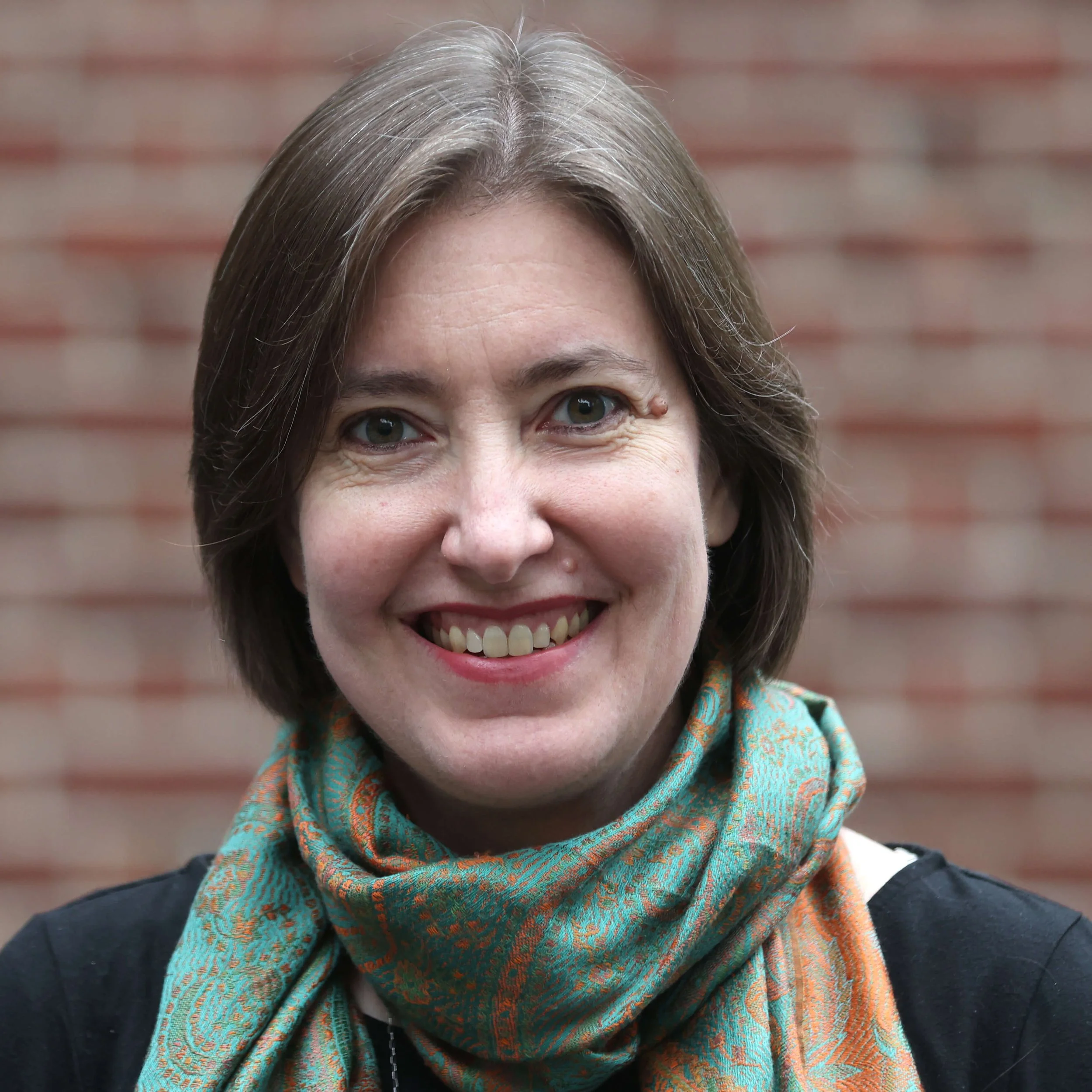 Close-up of a woman with shoulder-length gray hair, green eyes, and a warm smile, wearing a colorful patterned scarf, standing against a red brick wall.