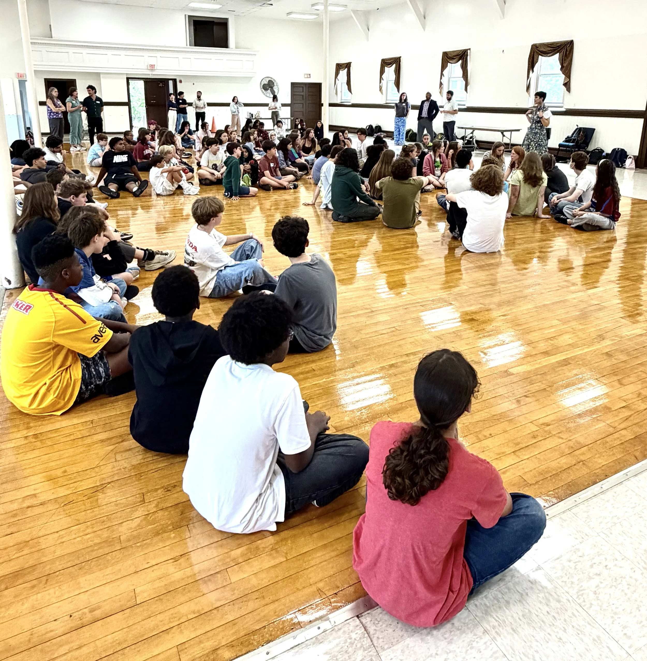 Large group of students sitting in a hall, listening to a teacher.