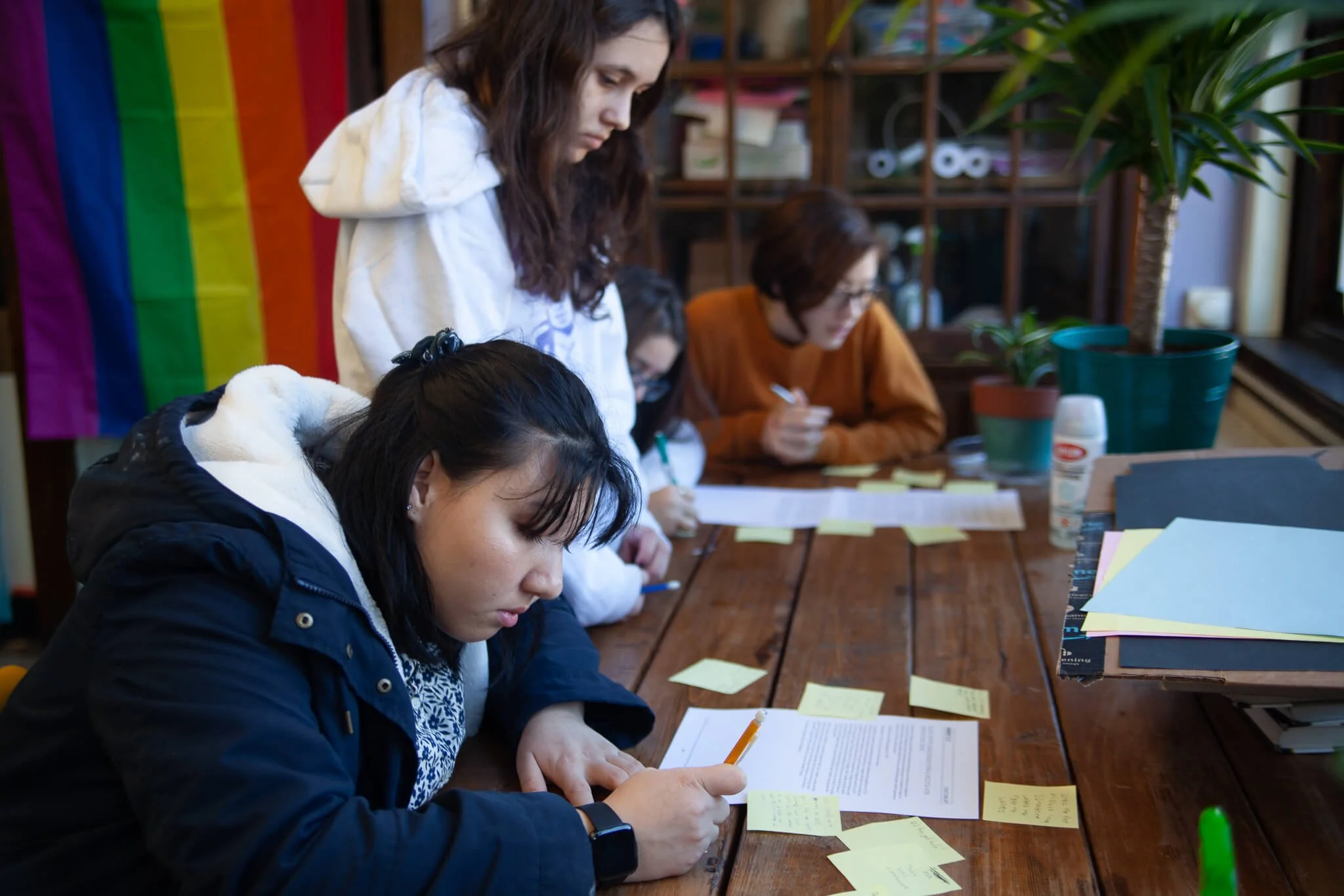 Several people working at a table with papers, sticky notes, and writing tools in a cozy indoor space, with a rainbow pride flag and plants visible in the background.