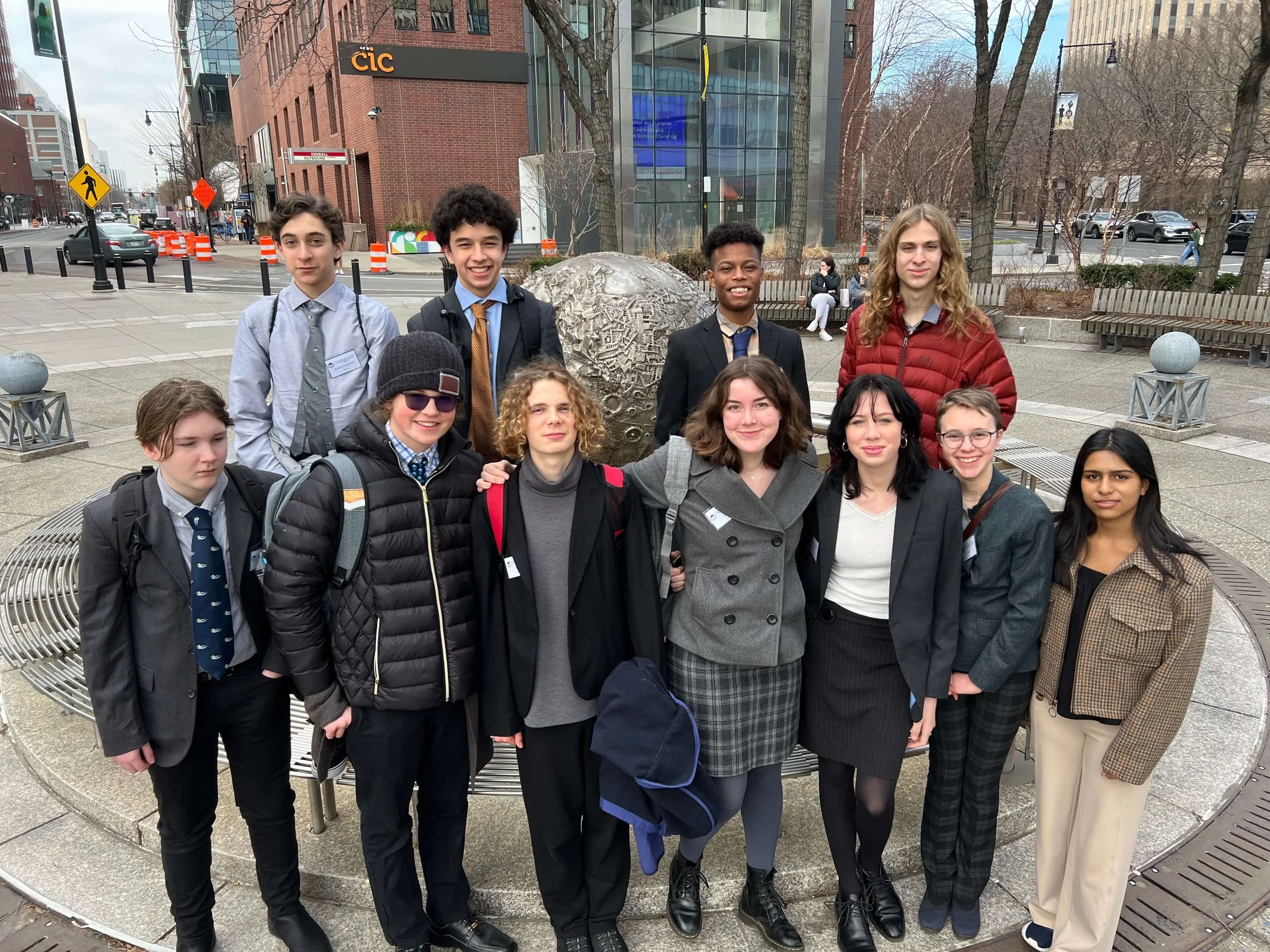 A group of diverse teenagers standing together outdoors in an urban setting with brick and glass buildings in the background. They are smiling and dressed in casual and semi-formal clothing, with some wearing backpacks and jackets. There are benches and trees around them.