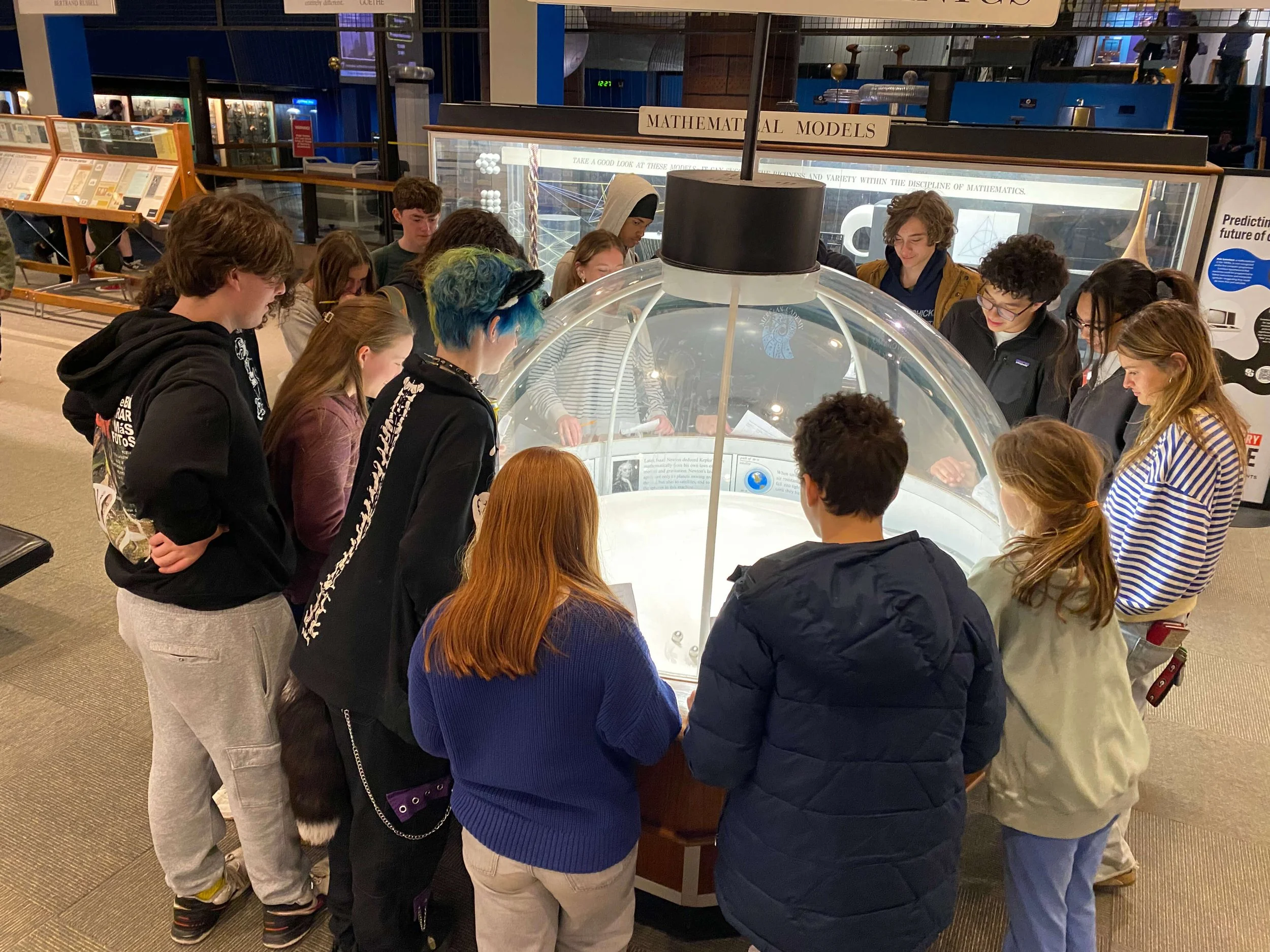 A group of people gathered around a science exhibit at a museum, observing a display inside a glass dome labeled 'Mathematical Models'.