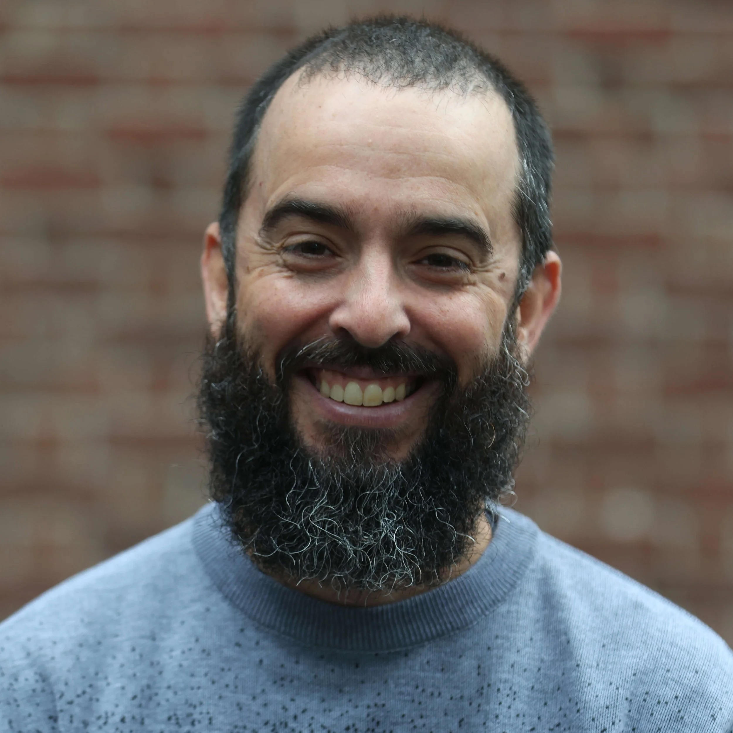 Close-up photo of a man's face, showing a partially shaved head, beard, and smiling expression, with a blurred brick wall background.