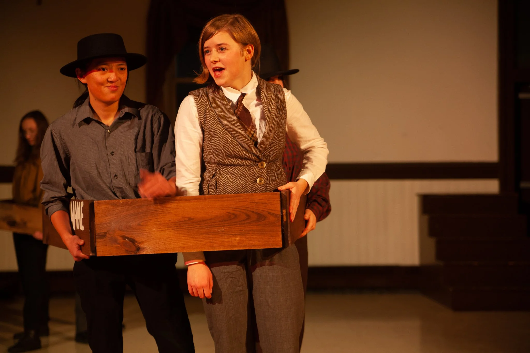 People dressed in vintage clothing participating in a theatrical performance, holding a wooden box, with a stage setting in the background.