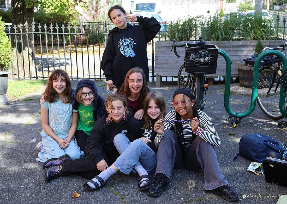 Group of seven young girls sitting and standing outdoors on pavement, smiling, with bicycles and greenery in the background.