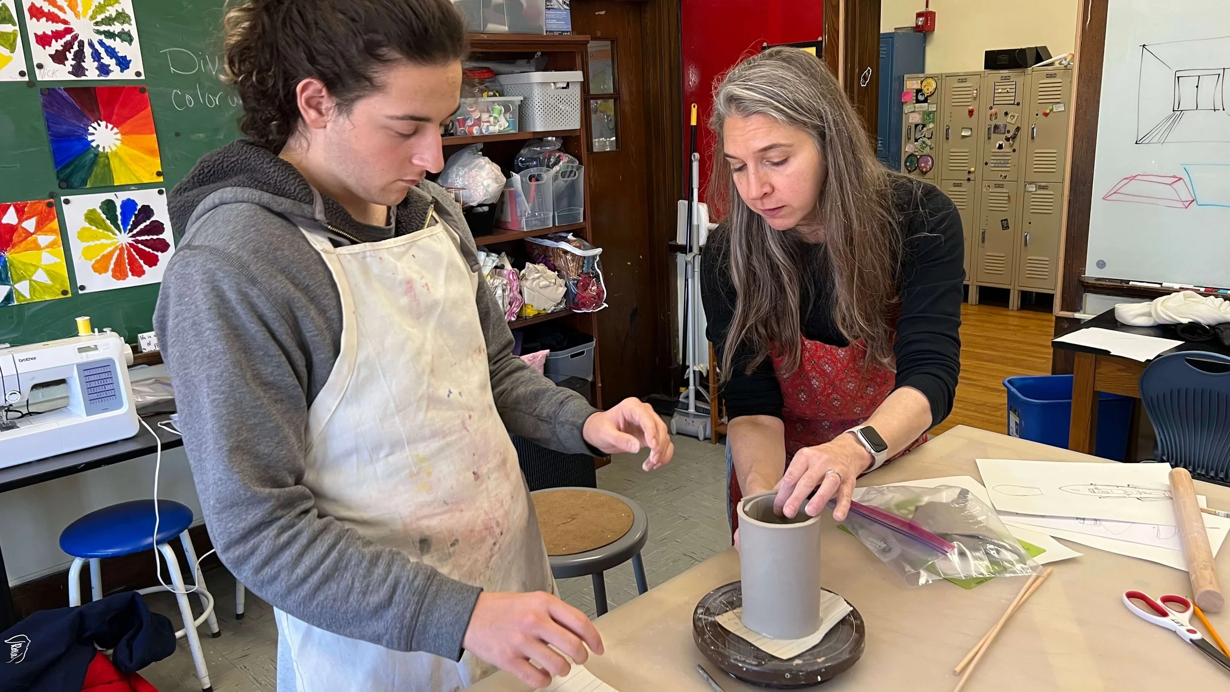 A woman and a teenage boy working on a craft project at a classroom table, with art supplies and sketches around them.