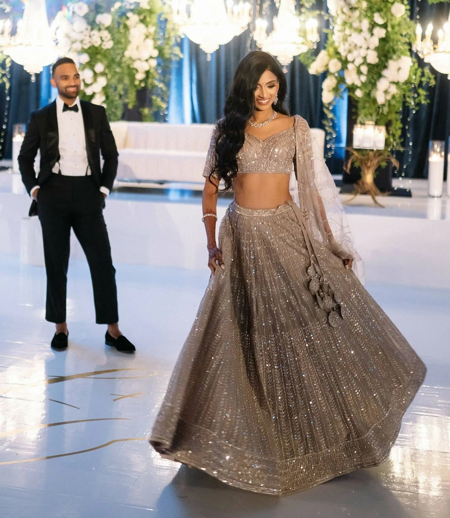A woman in a gold, glittery lehenga with a matching choli and dupatta, smiling and looking down, at a decorated event, alongside a man in a black tuxedo with a bow tie, on a dance floor with floral arrangements and chandeliers in the background.