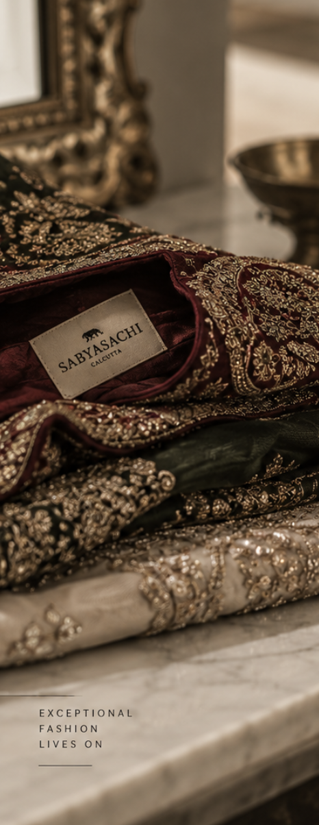 Stacked embroidered and embellished fabrics on a marble surface, with a label reading 'SABYASACHI CALCUTTA,' and a gold decorative bowl in the background.