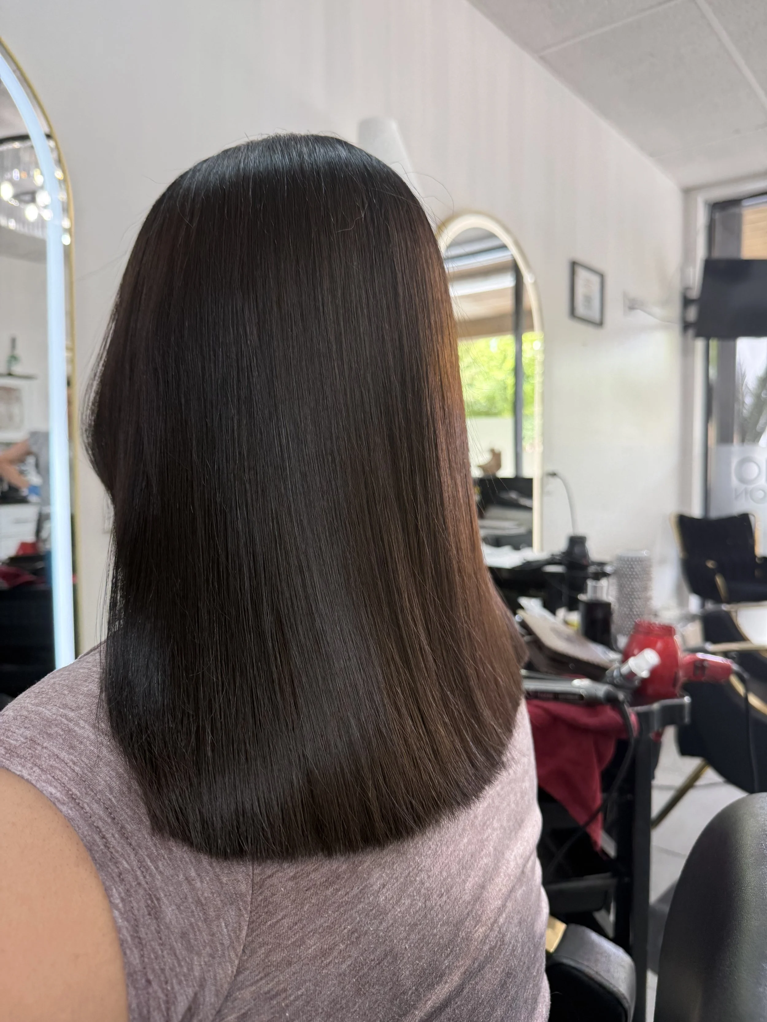 A woman with long, straight, dark brown hair sitting in a salon or hair studio, with hair styled smoothly and shiny. The background shows salon equipment and a mirror.