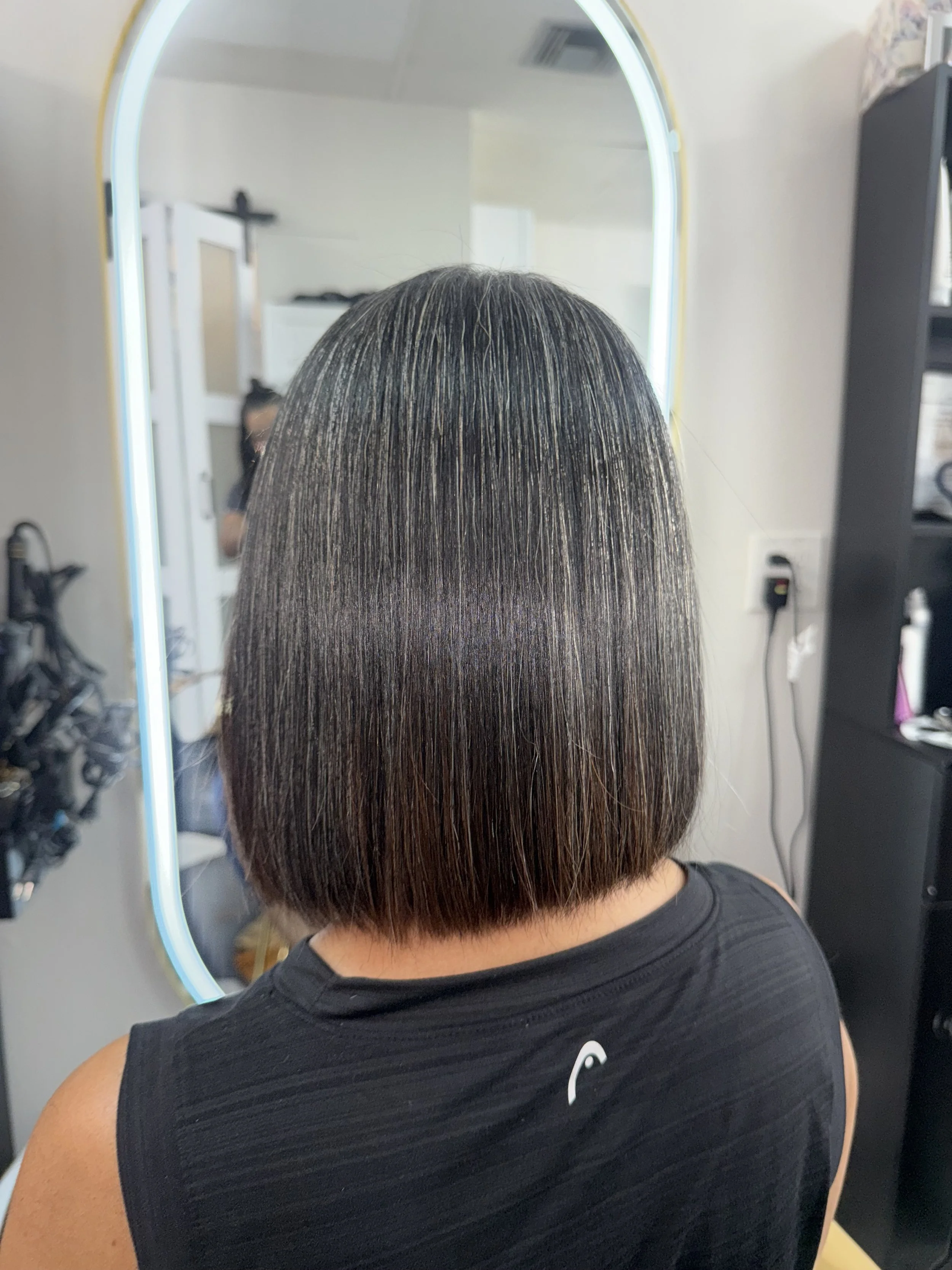 A person with straight, shoulder-length dark hair with some light streaks, facing away in front of a mirror in a hair salon.