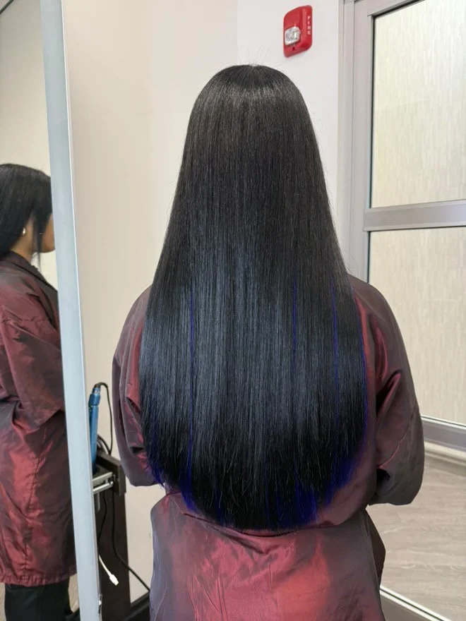 A woman with long, straight, shiny black hair with blue highlights, standing in front of a mirror in a salon.