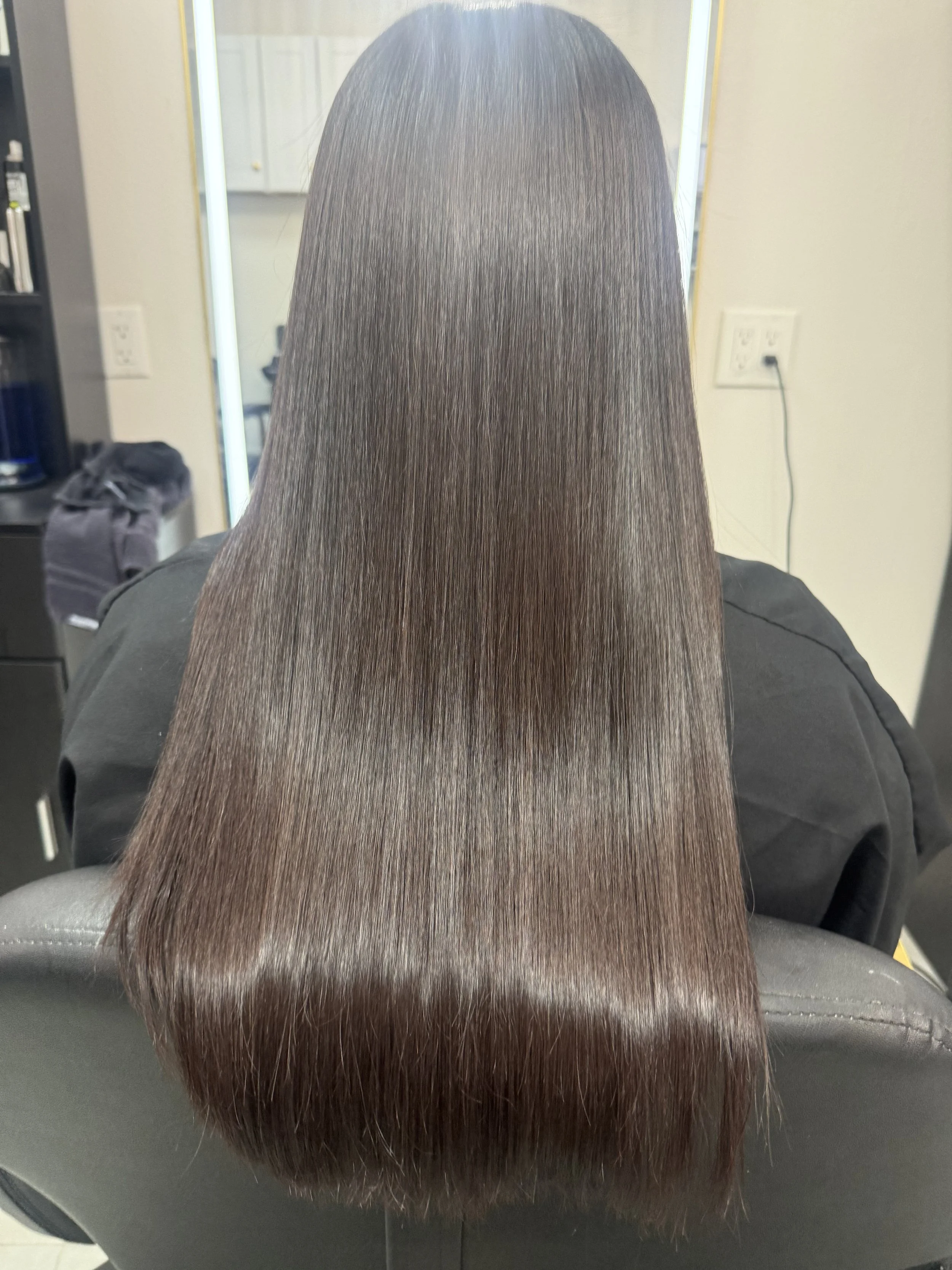 A person with long, straight, shiny brown hair sitting in a salon chair, facing a mirror.
