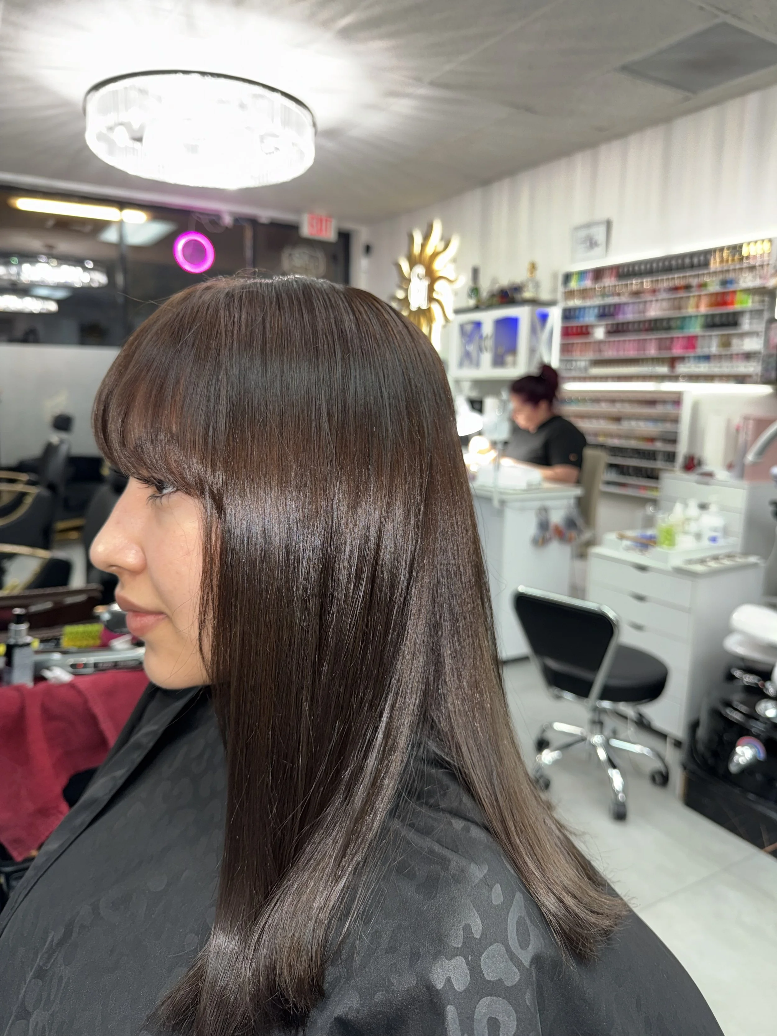 A woman with shiny, straight brown hair sitting in a salon, with shelves of nail polish and a stylist working in the background.