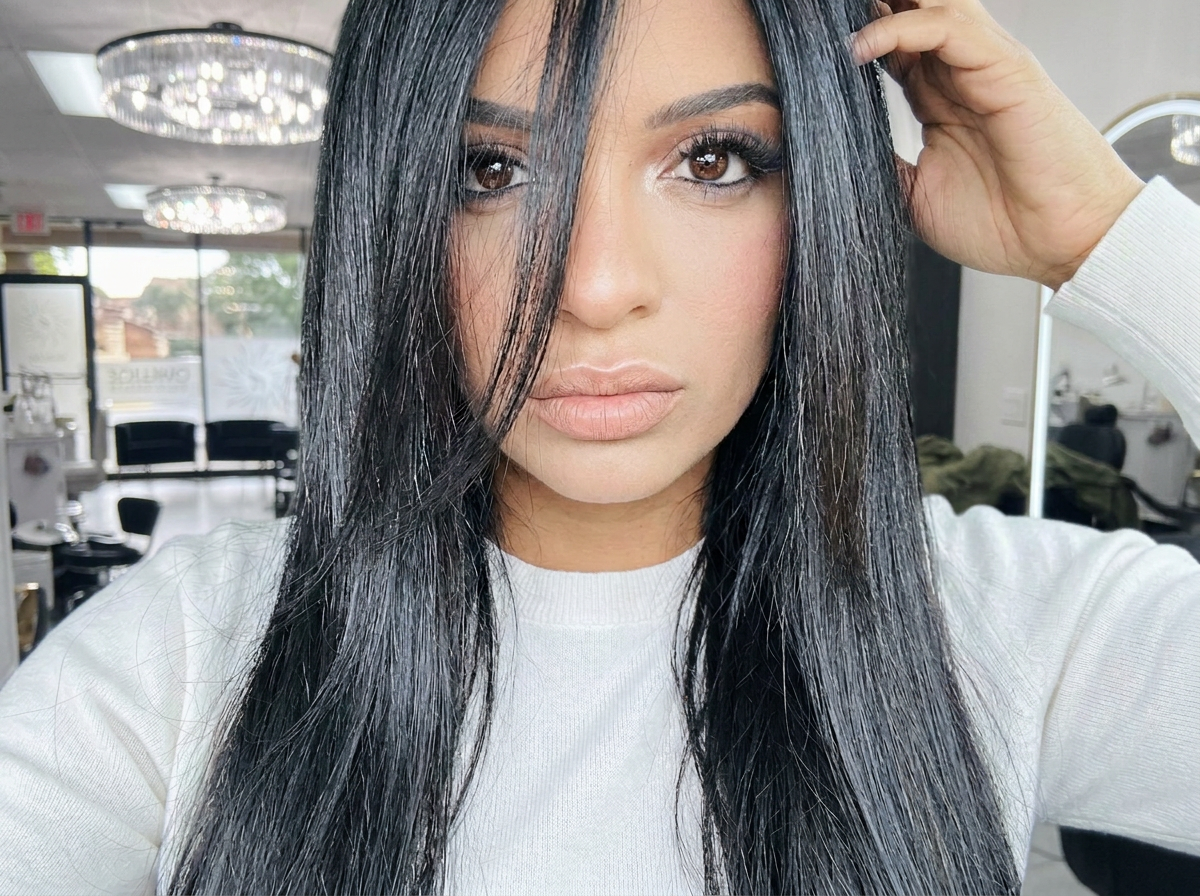 A woman with long black hair and brown eyes taking a selfie inside a well-lit salon or waiting area, with chandeliers and chairs visible in the background.