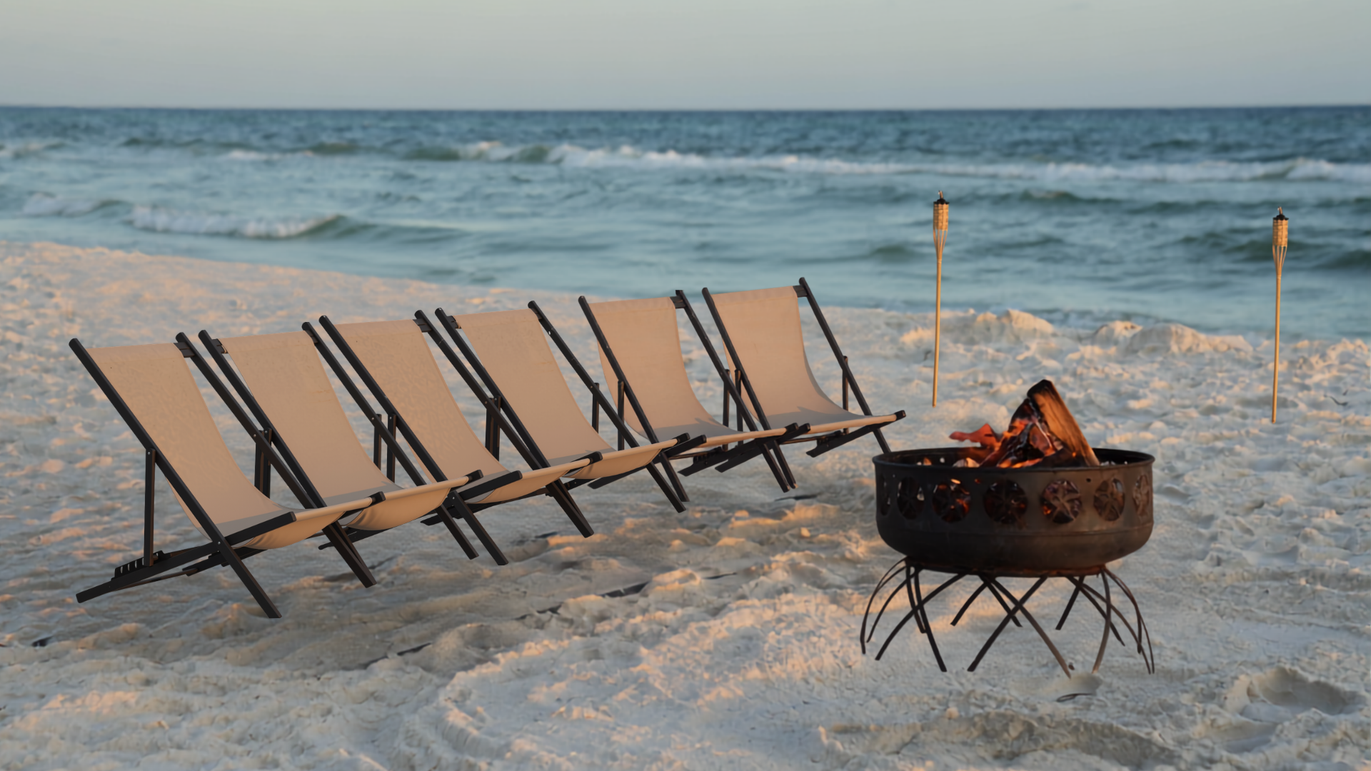 Beach scene with six empty lounge chairs facing the ocean, a fire pit with burning logs, two tiki torches, and the waves in the background during sunset.