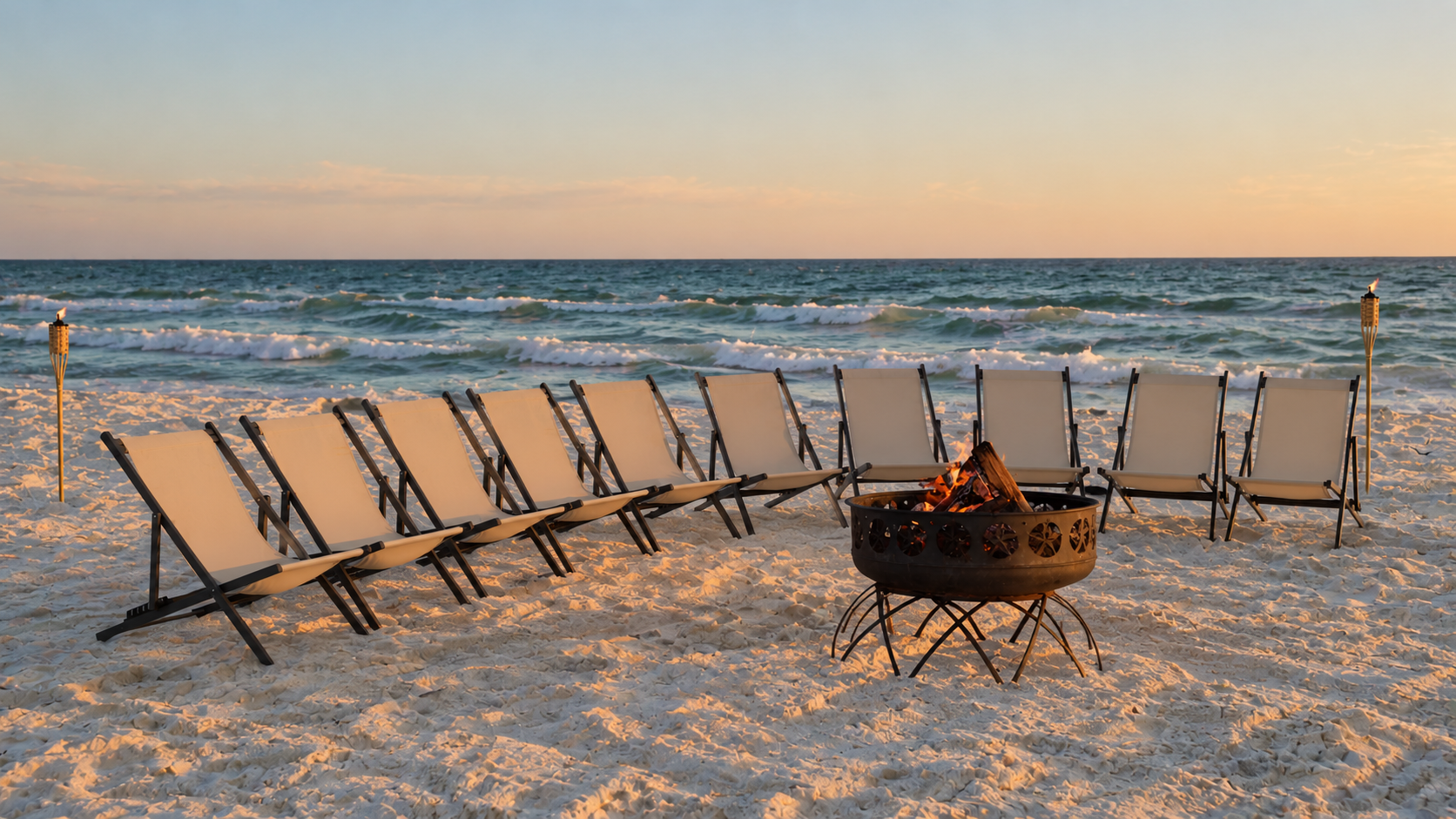 Beach scene with a row of empty chairs facing the ocean, a fire pit in front, and tiki torches on both sides, under a sunset sky.