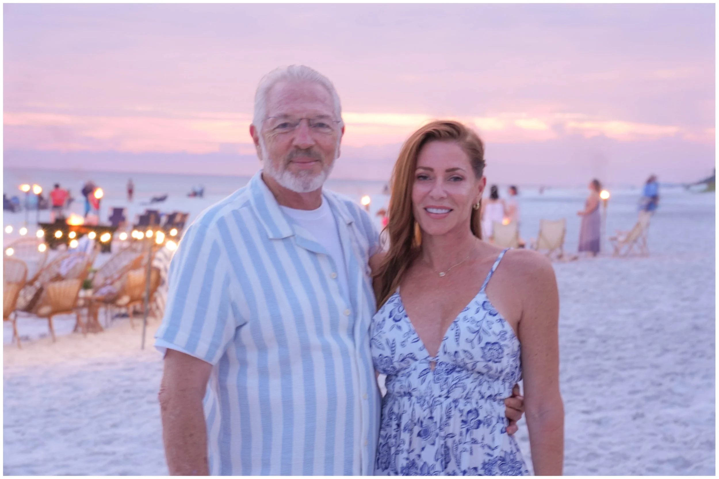 A man and woman standing on a beach at sunset, smiling and posing for a photo. The beach is decorated with chairs and lights, with several people in the background.