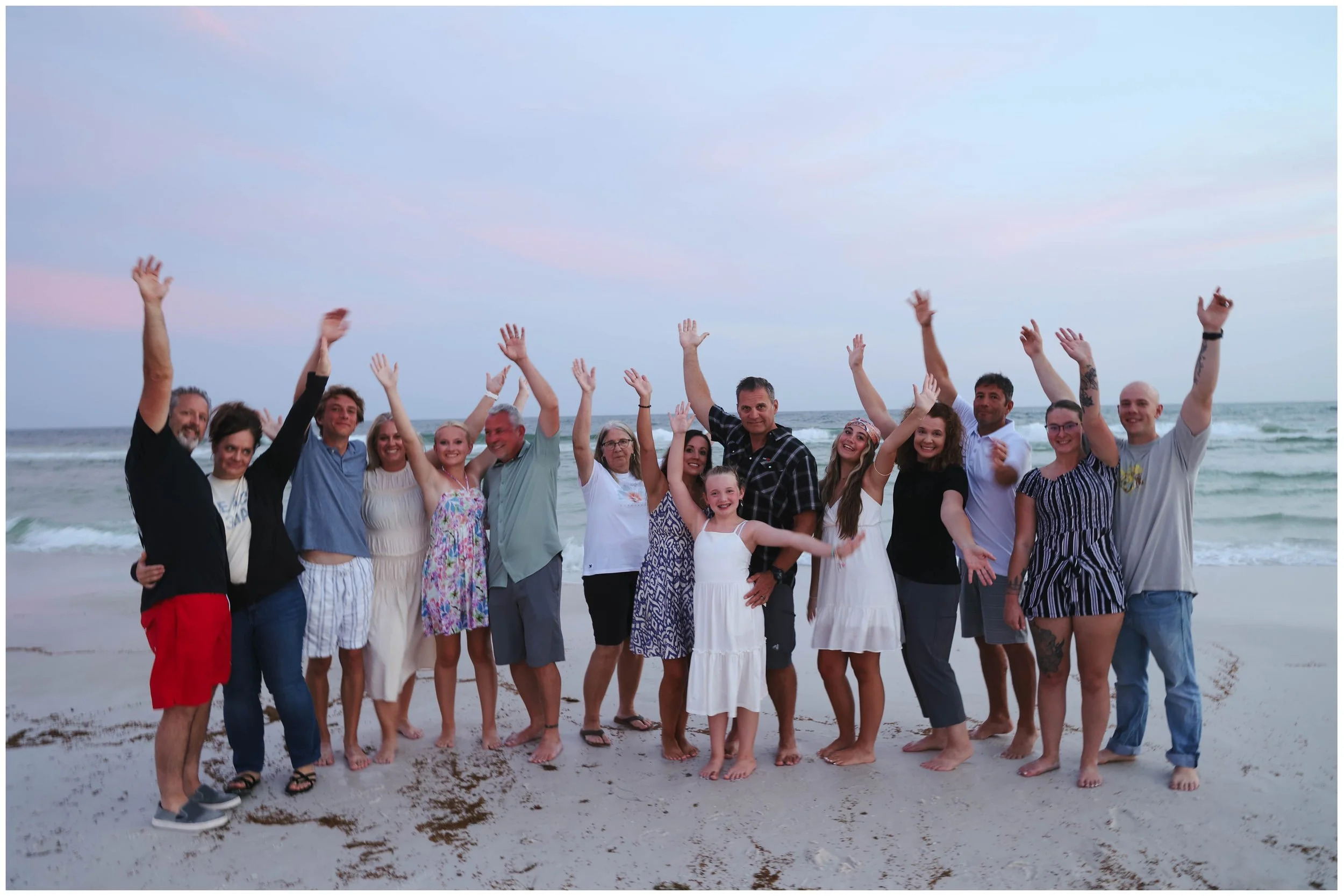 A group of diverse people, including men, women, and children, standing on a beach with waves in the background, raising their hands and smiling.