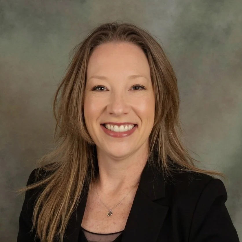 Portrait of a woman smiling, with long light brown hair, wearing a black blazer and a silver necklace, against a neutral background.
