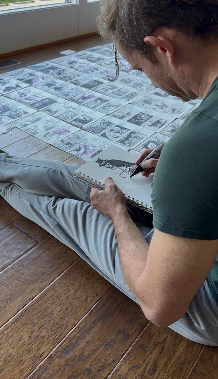 Man sitting on wooden floor, drawing a comic character in a sketchbook while looking at a collection of comic pages laid out on the floor.