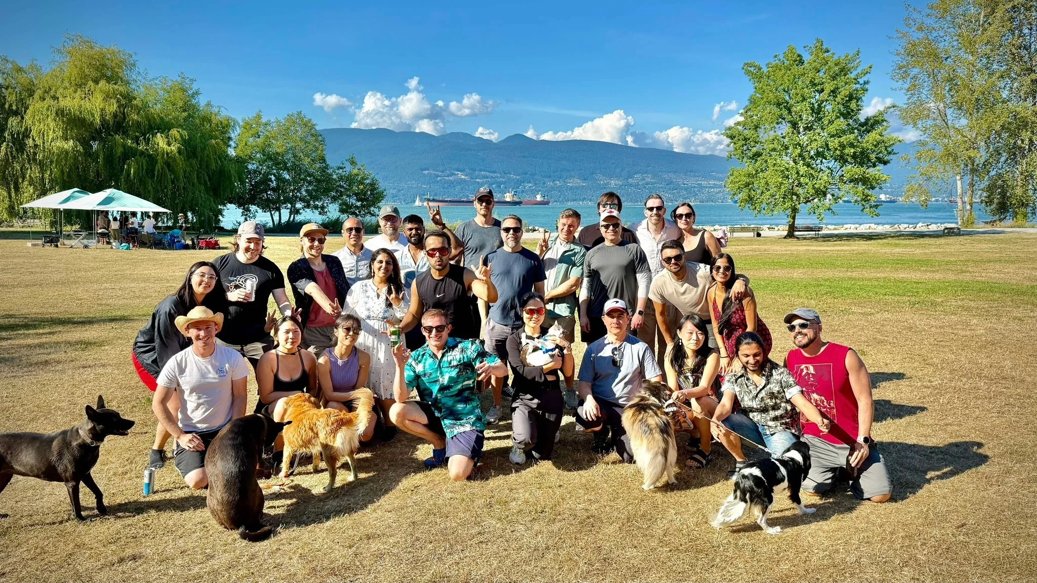 A large group of people posing for a photo outdoors in a park with trees, grass, and a body of water with mountains in the background. Some people are holding dogs and wearing sunglasses, hats, and casual summer clothing.