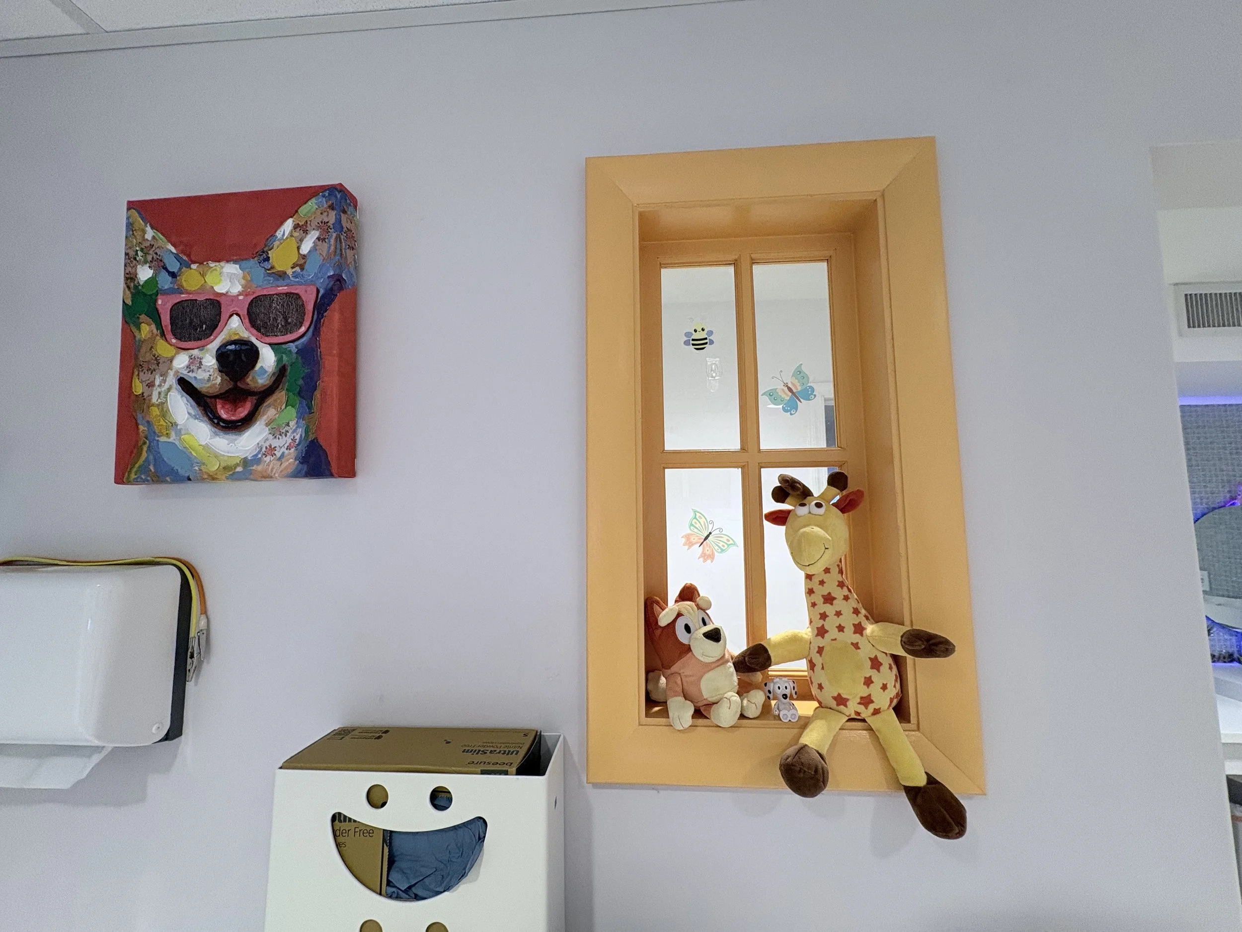 Colorful painting of a dog wearing pink sunglasses hanging on a white wall in a room. Below a yellow window frame with various stuffed animals, including a large giraffe plush toy with a long neck, smaller plush toys, and decorative butterfly sticker