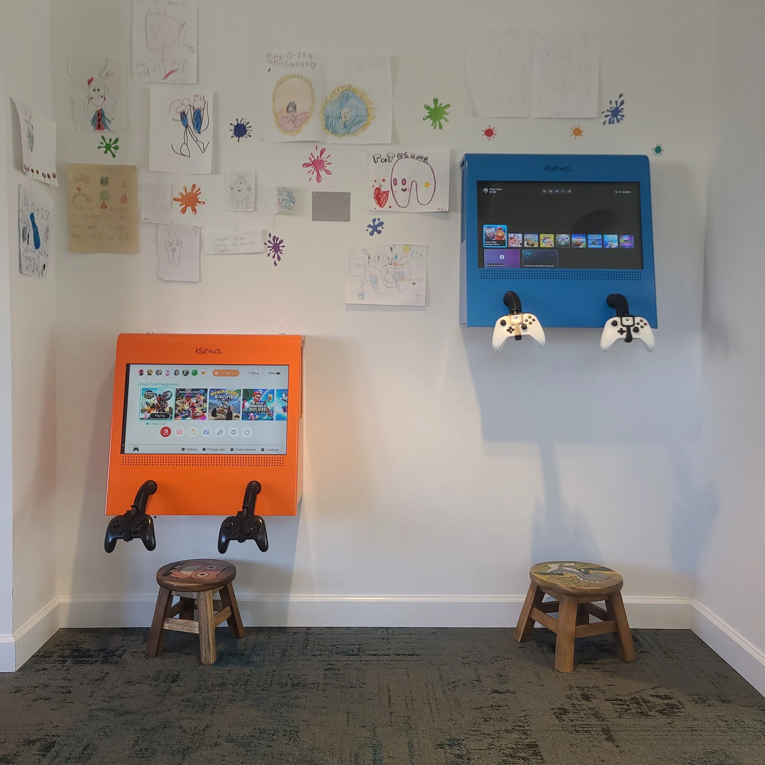 A wall with children's drawings and colorful splash decorations above a gaming kiosk and two wooden stools.