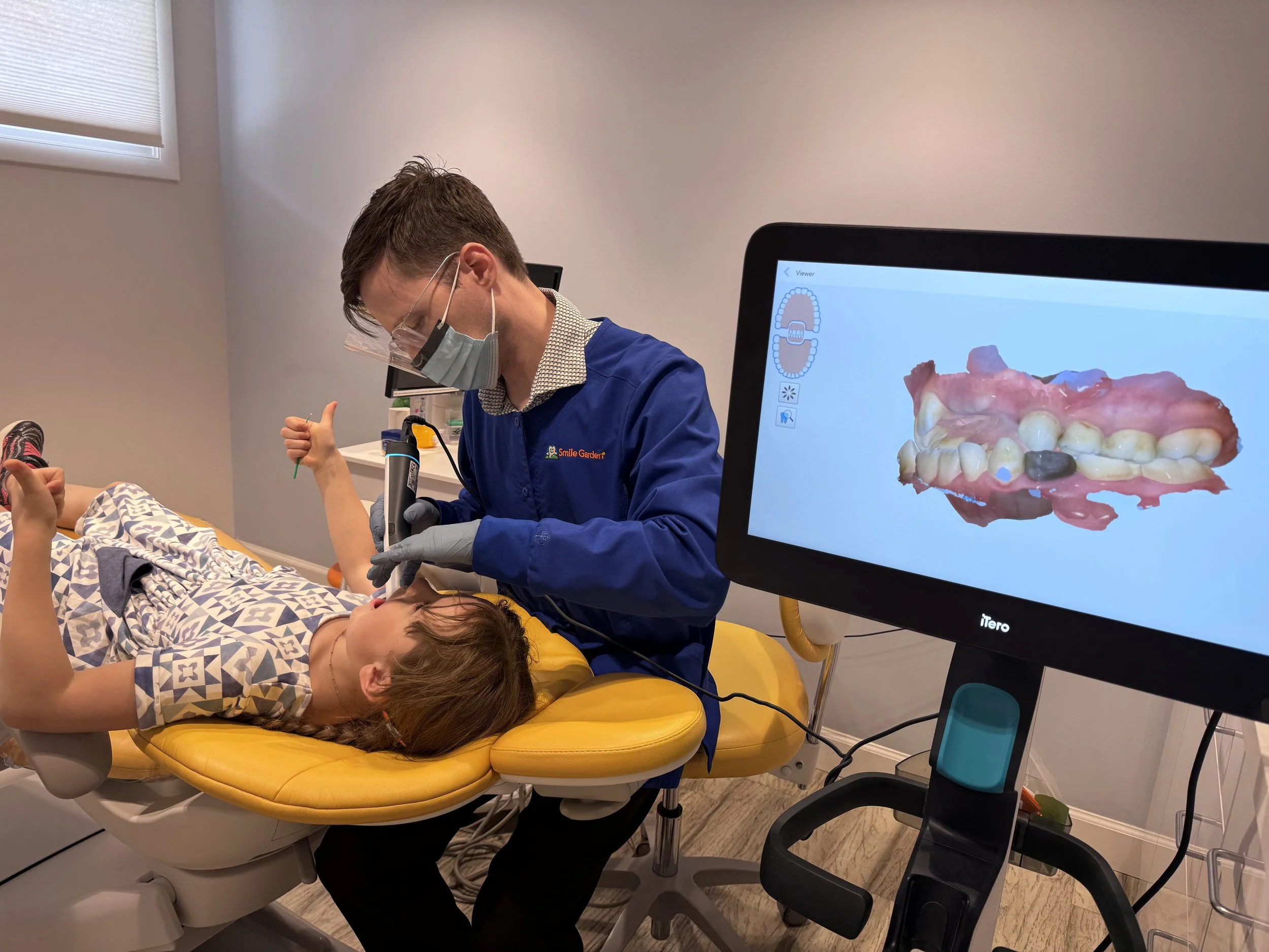 Dentist wearing protective gear examining a patient's teeth using a dental instrument in a clinic, with digital dental scan displayed on a monitor in the foreground.
