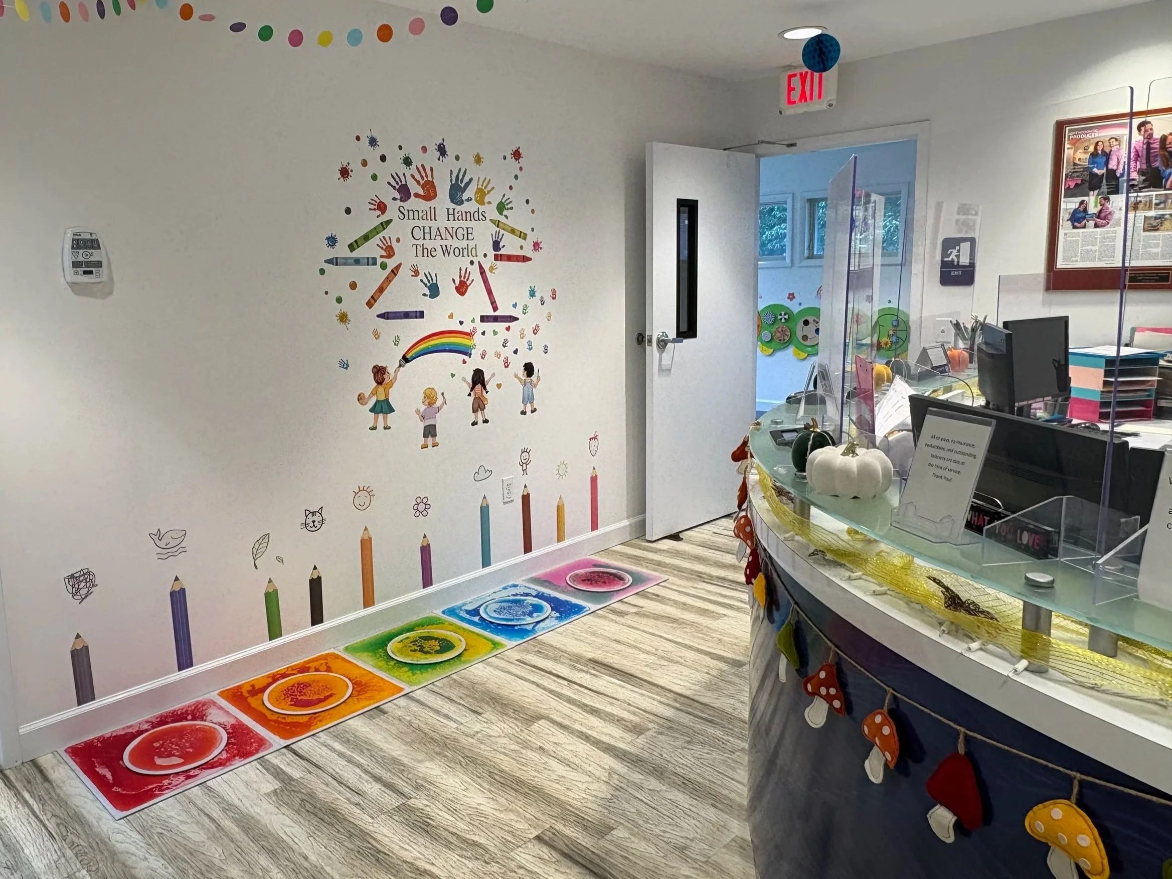 Colorful chalk drawings on the wall depicting children, paint cans, and the phrase 'Small Hands CHANGE The World.' A rainbow, small doodles, and rainbow-colored paint palettes are part of the mural. A rainbow-colored paint tray with circular paint we